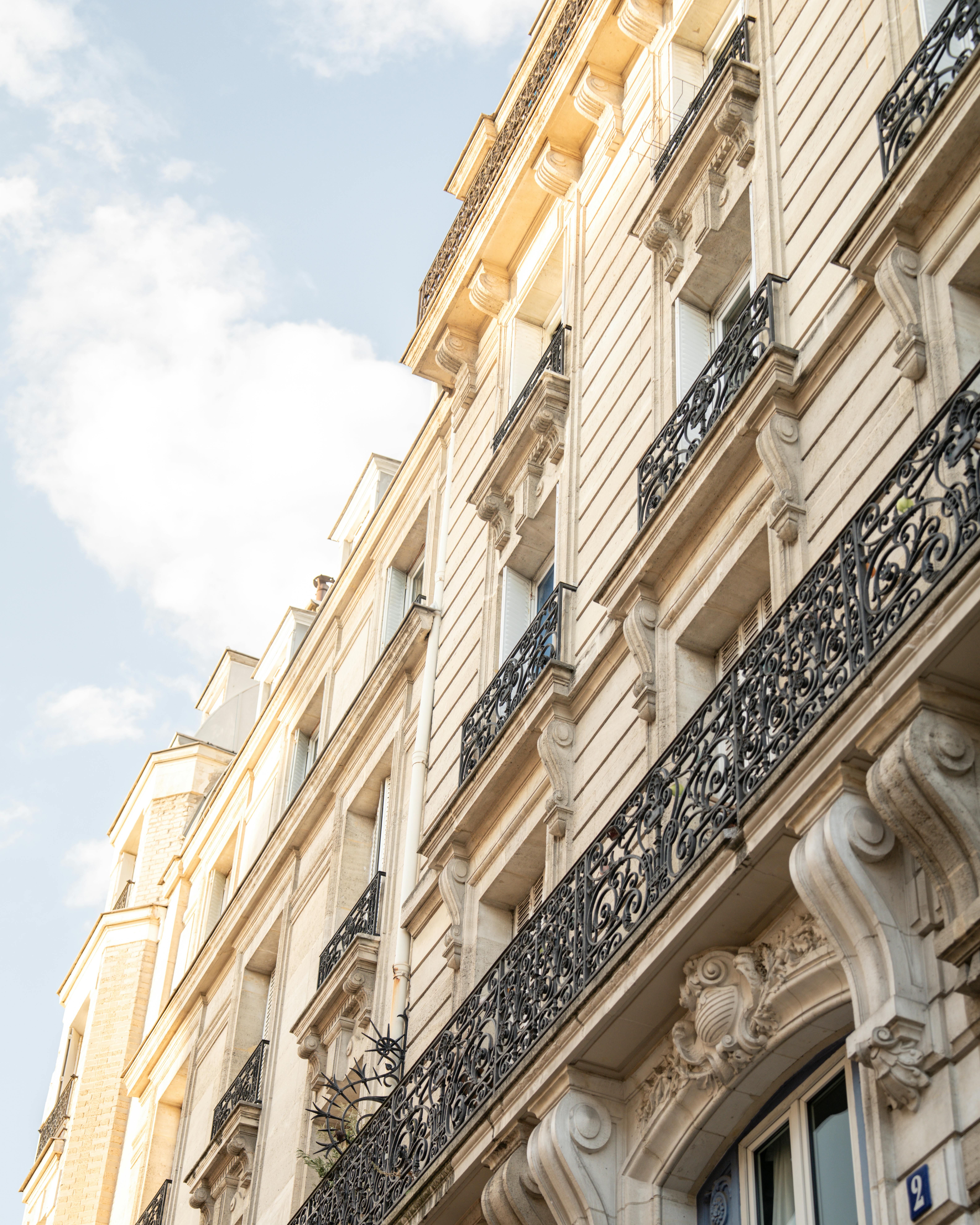 Ornate Parisian-style beige stone building facade with black wrought iron balconies under a partly cloudy sky.