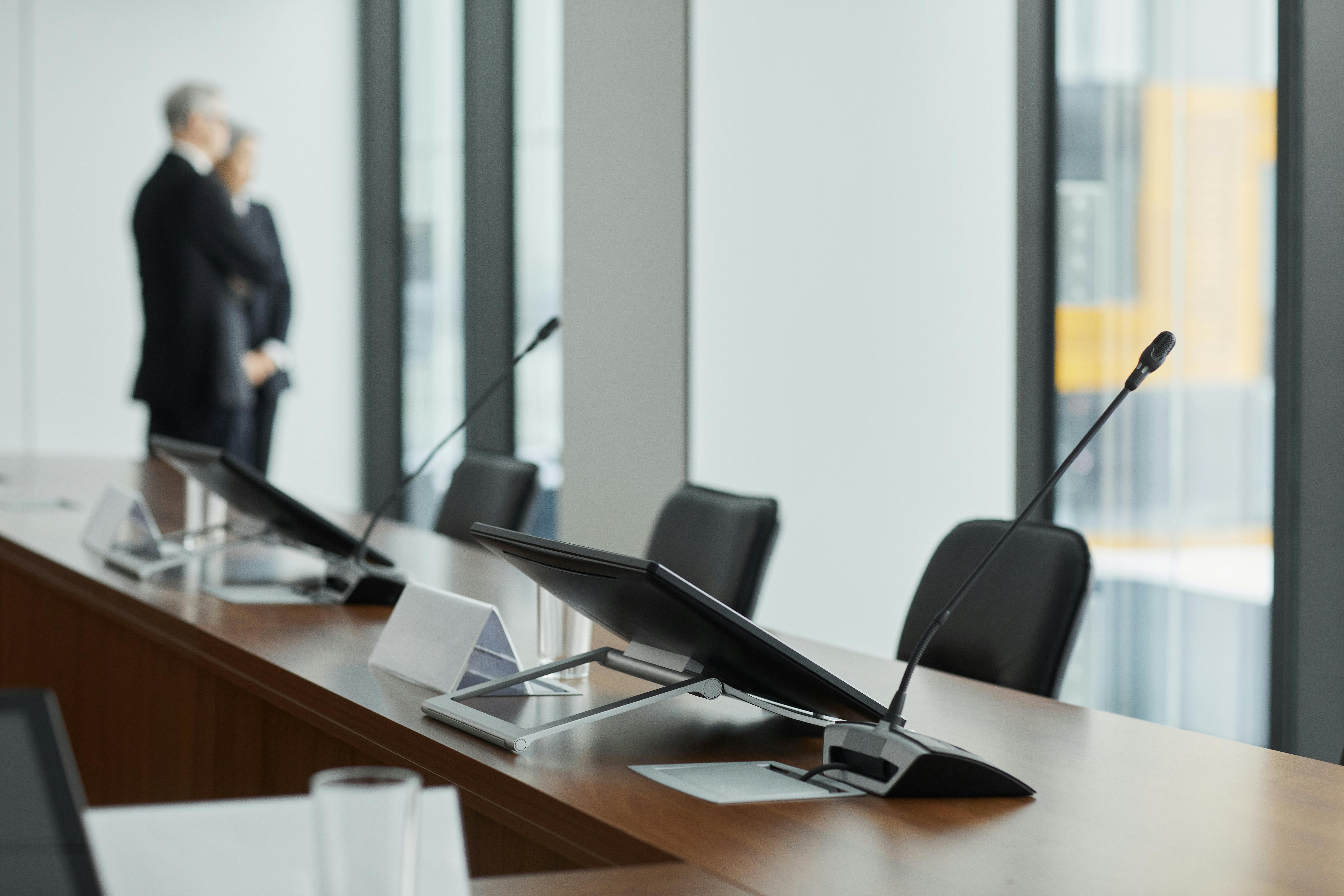 Conference room table with microphones and angled monitors, two people blurred in background.