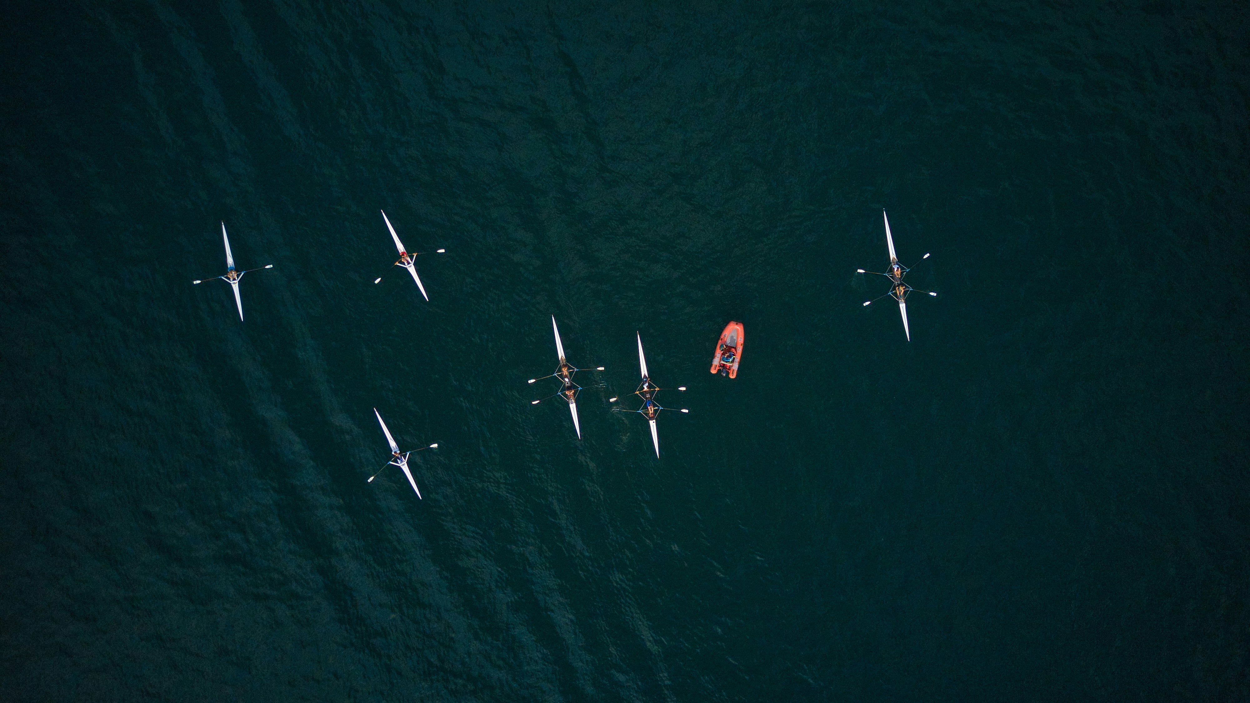 Aerial view of six rowing teams on dark water with a small orange motorboat nearby.