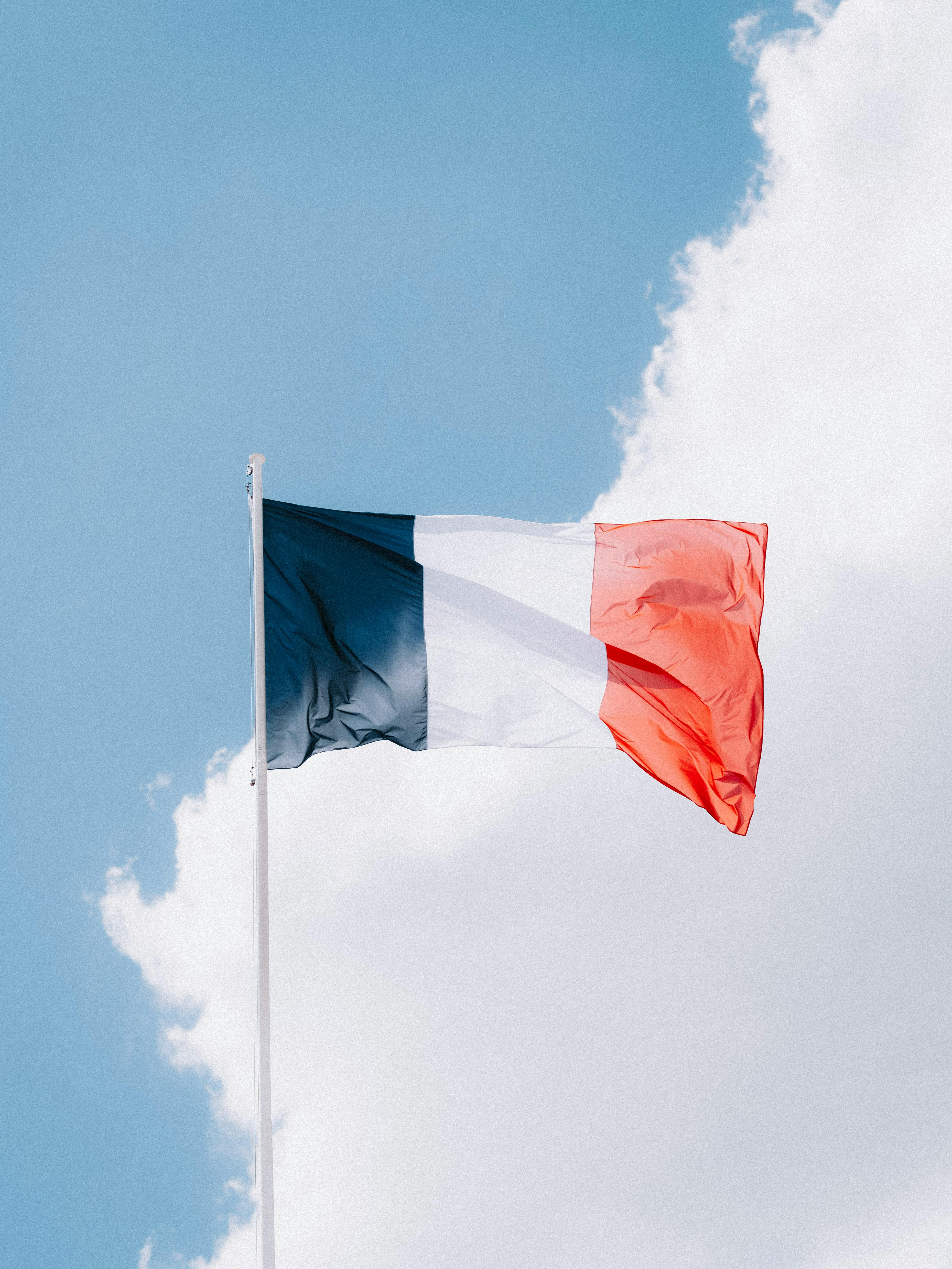 French flag with blue, white, and red vertical stripes flying against a partly cloudy sky.