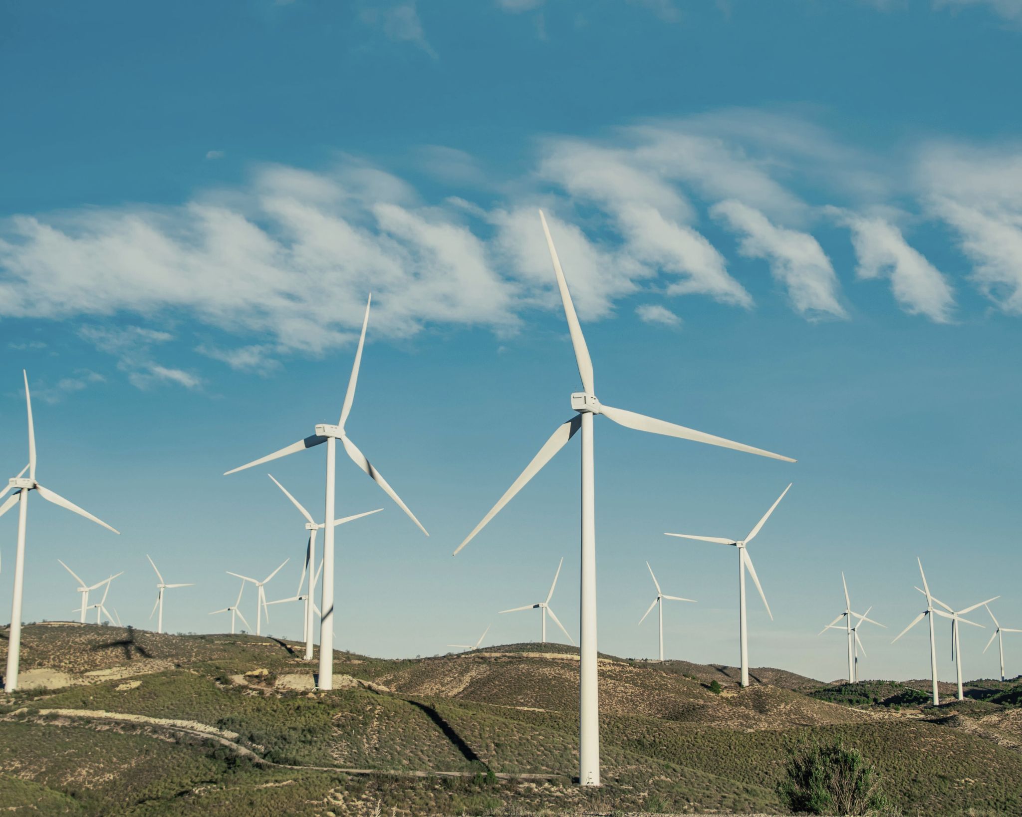 Multiple white wind turbines on green hills under a blue sky with scattered clouds.