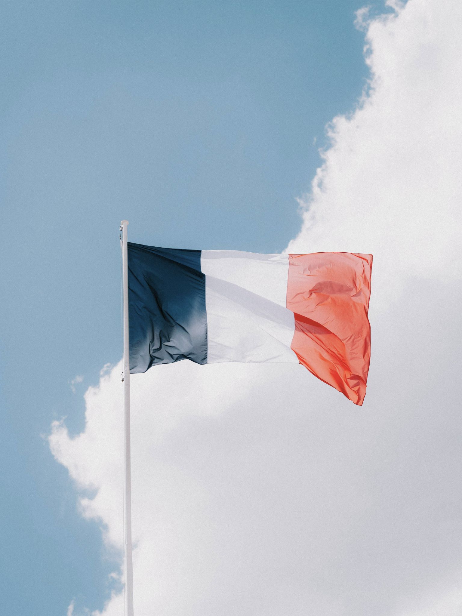 French flag with blue, white, and red vertical stripes waving against a partly cloudy sky.