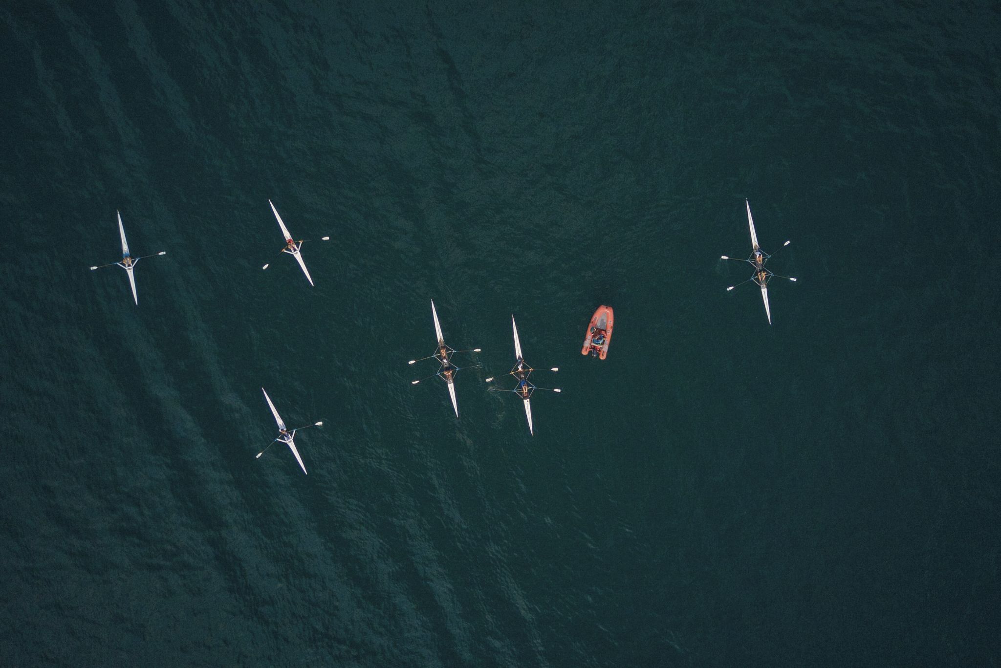 Aerial view of rowers in single and double scull boats on dark water with a small orange motorboat nearby.
