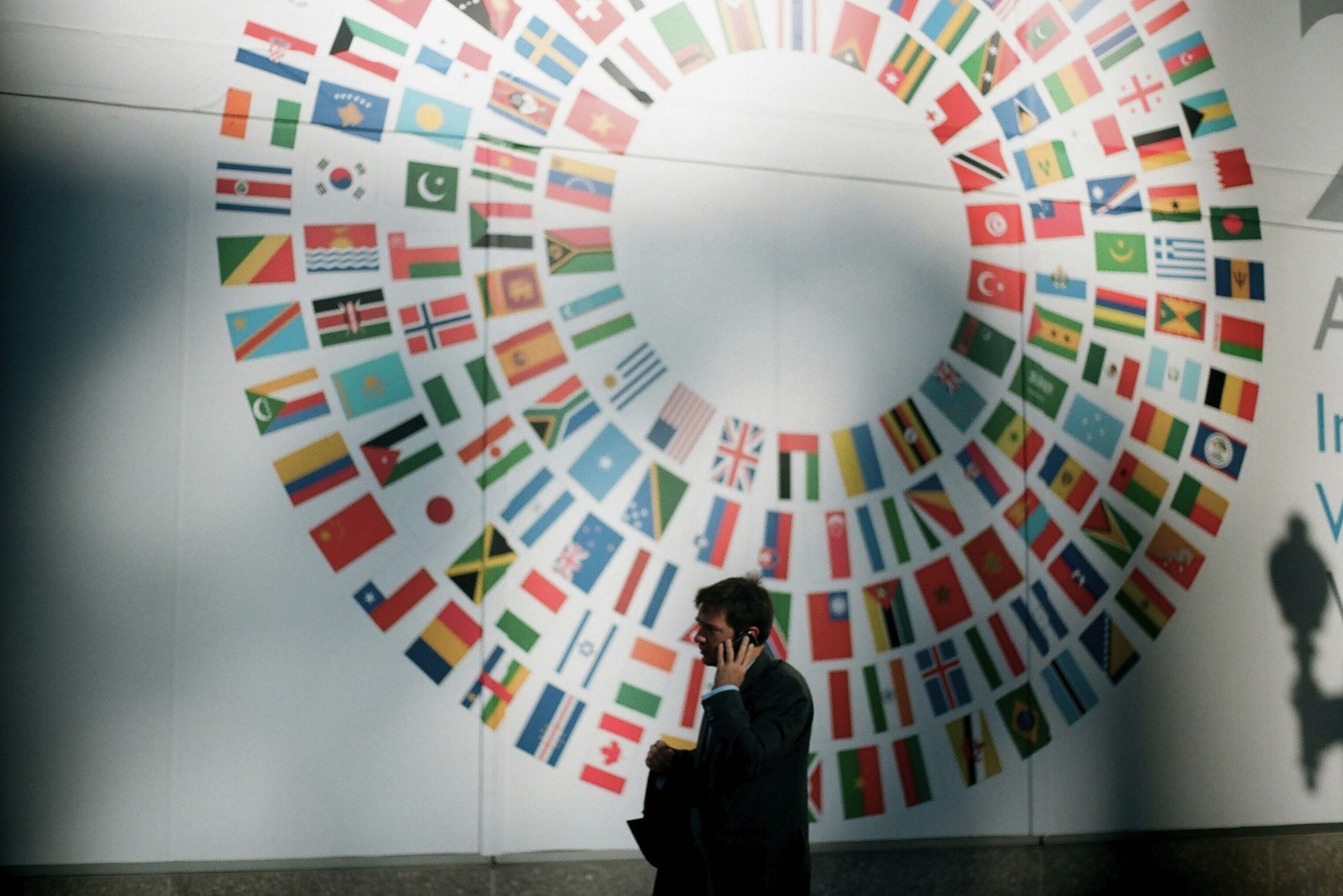 Man in business attire talking on a mobile phone walking in front of a large circular arrangement of international flags on a wall.