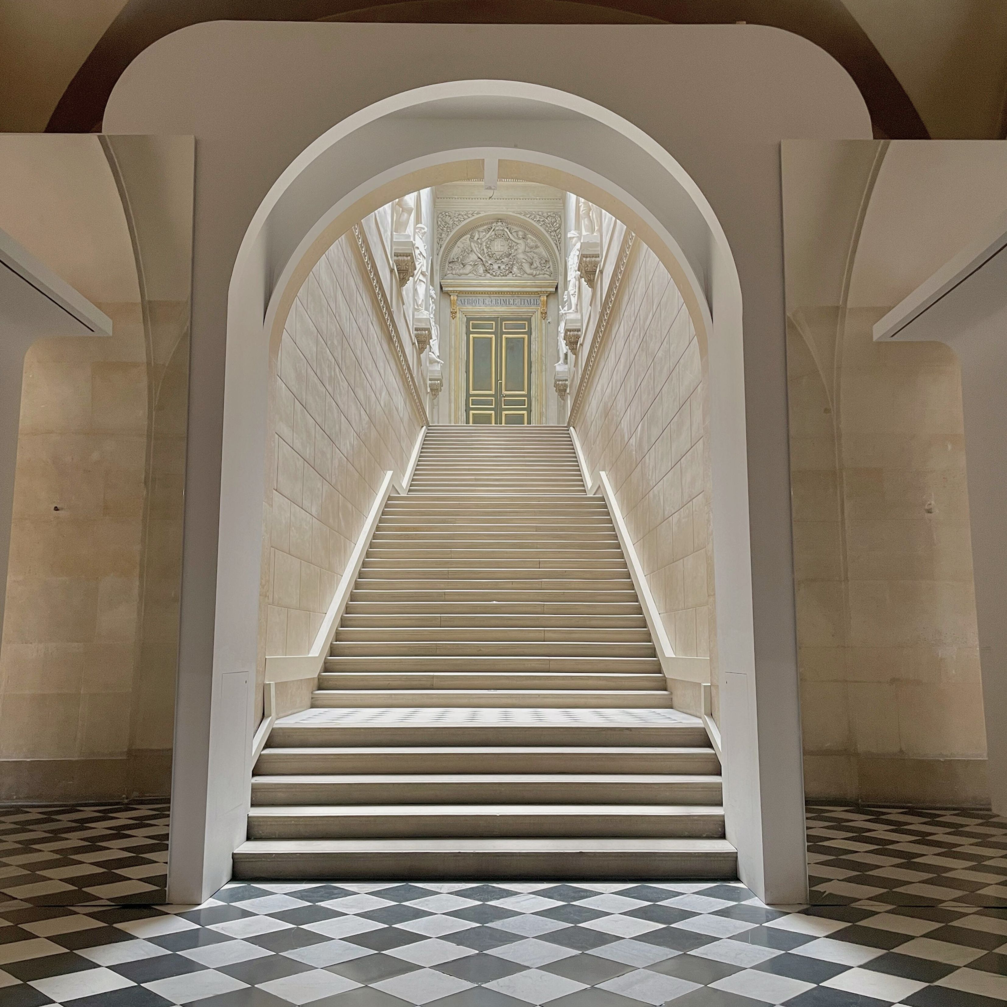 Symmetrical stone staircase with white walls leading up to a decorative green and gold door framed by sculptures, and a black and white checkered floor.