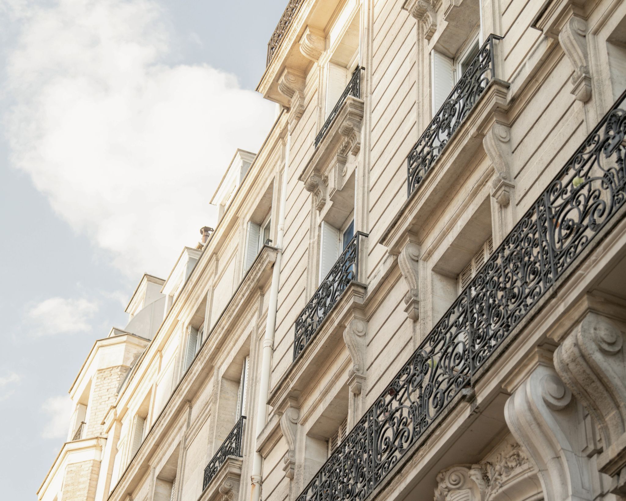 Light stone building facade with ornate black wrought iron balconies under a partly cloudy sky.