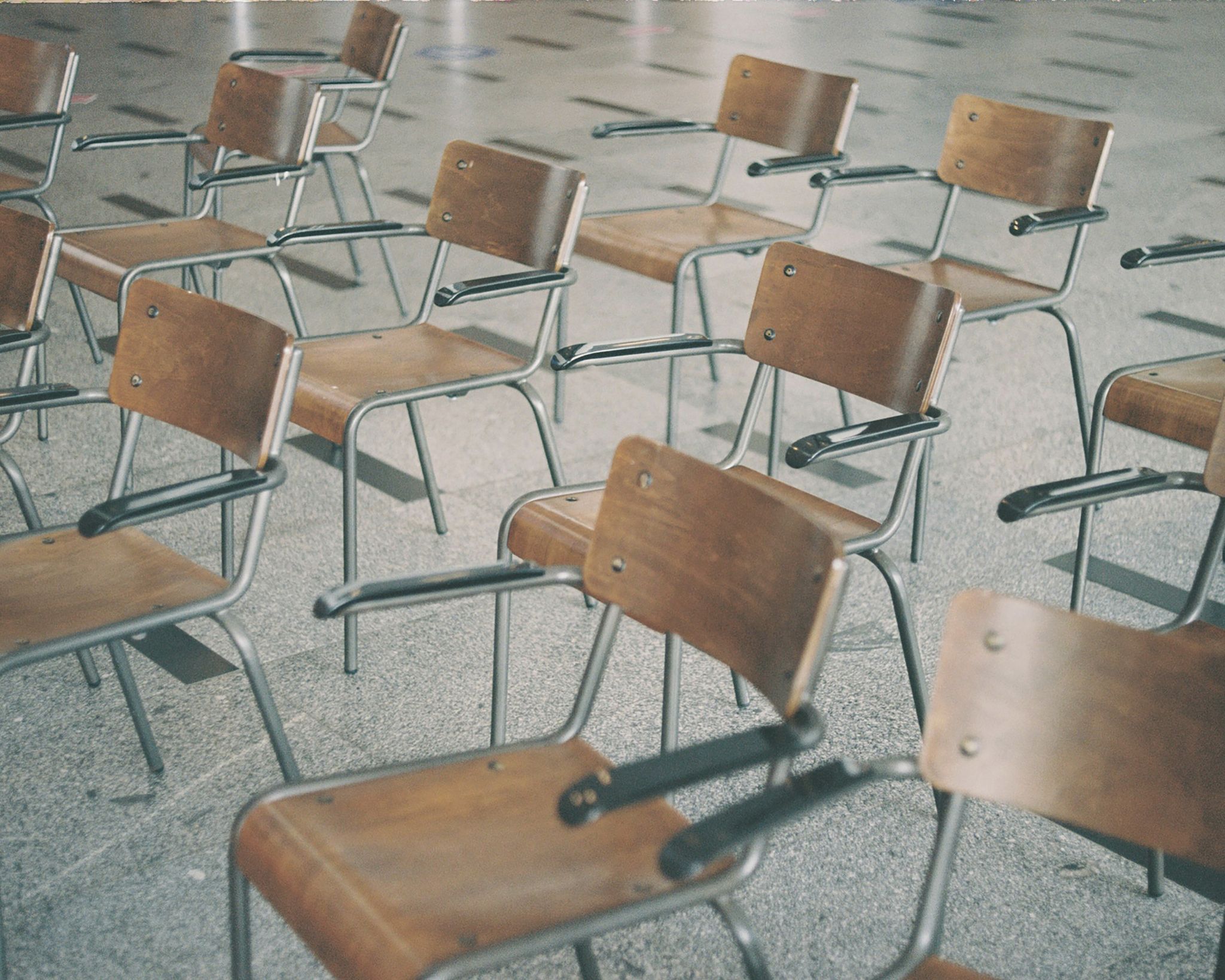 Empty classroom chairs with wooden seats and backrests and metal armrests arranged on a speckled floor.