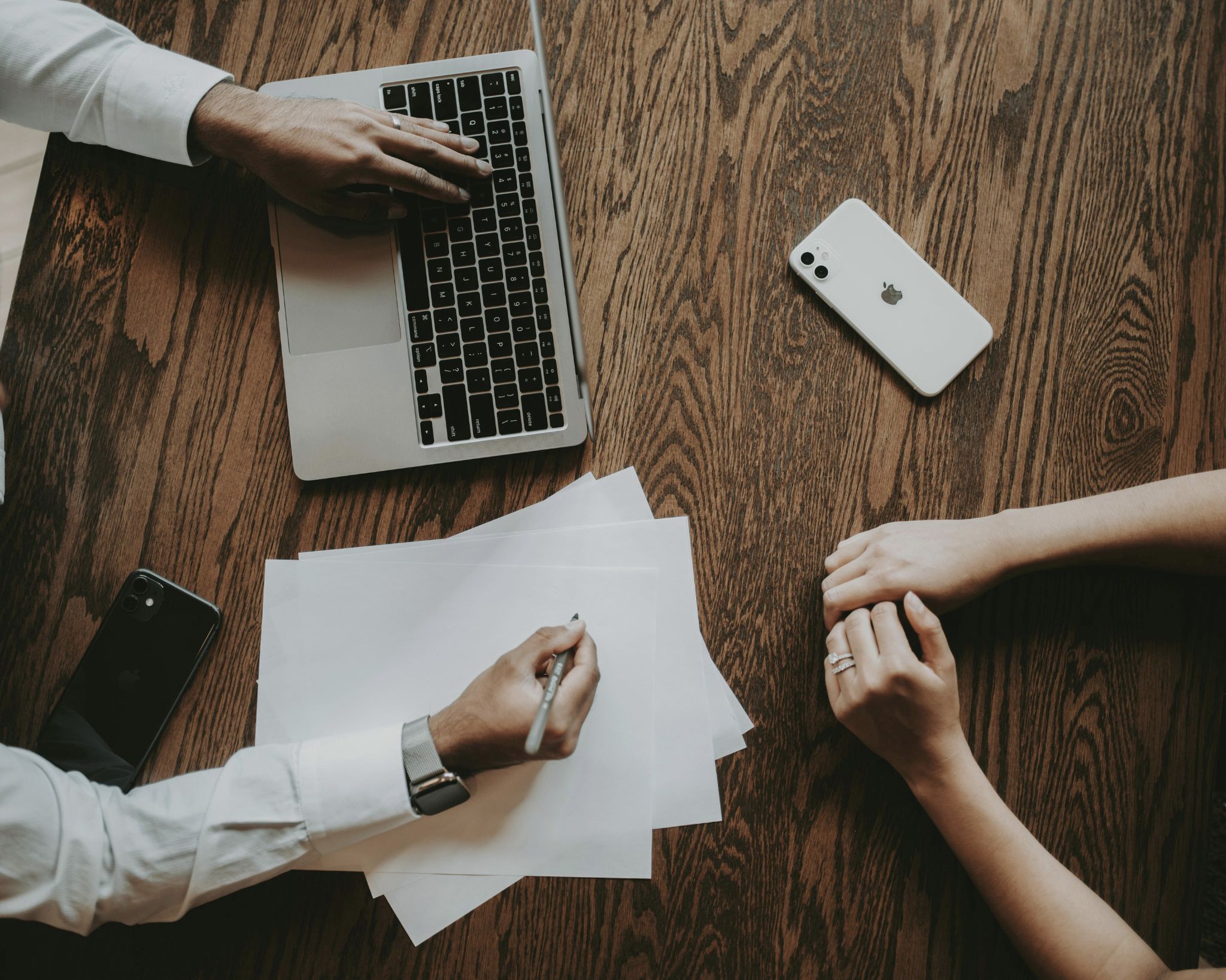 Two people at a wooden table, one writing on blank white papers with a pen near a laptop, the other resting hands nearby with a white iPhone on the table.