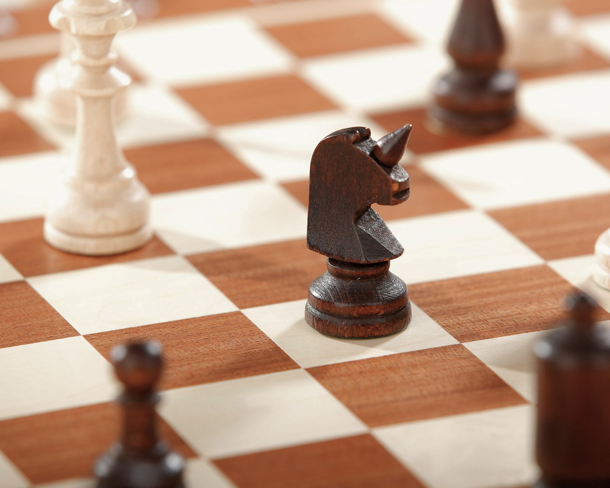 Close-up of a dark wooden knight chess piece shaped like a unicorn on a wooden chessboard with other chess pieces blurred in the background.