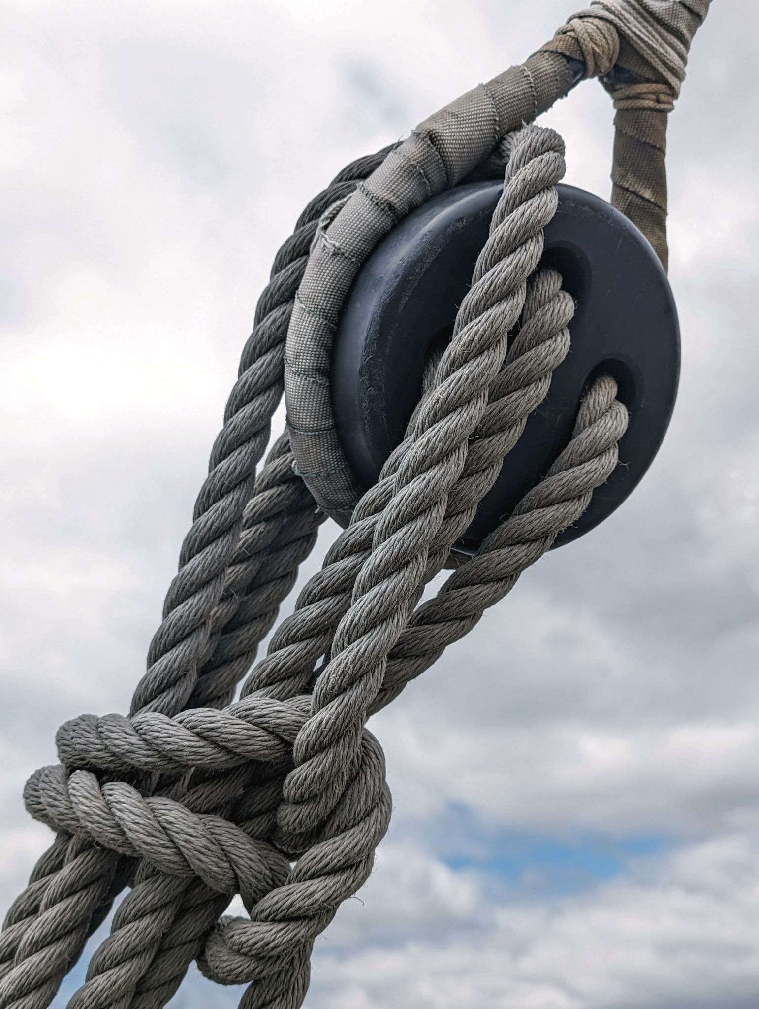 Close-up of thick gray ropes threaded through a black pulley against a cloudy sky.