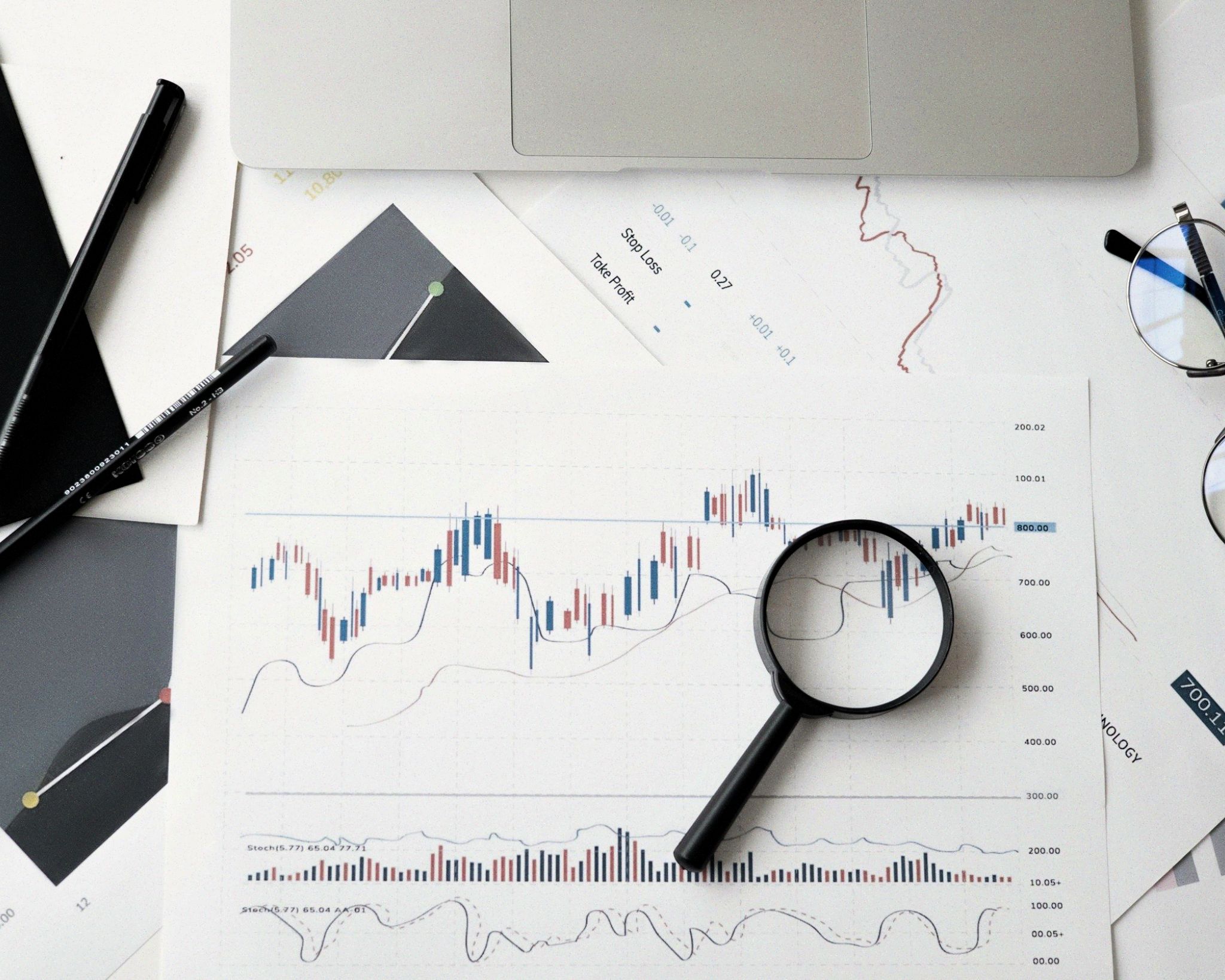 Magnifying glass on a financial chart with candlestick patterns and volume bars, surrounded by a laptop, glasses, and pens on a white desk.