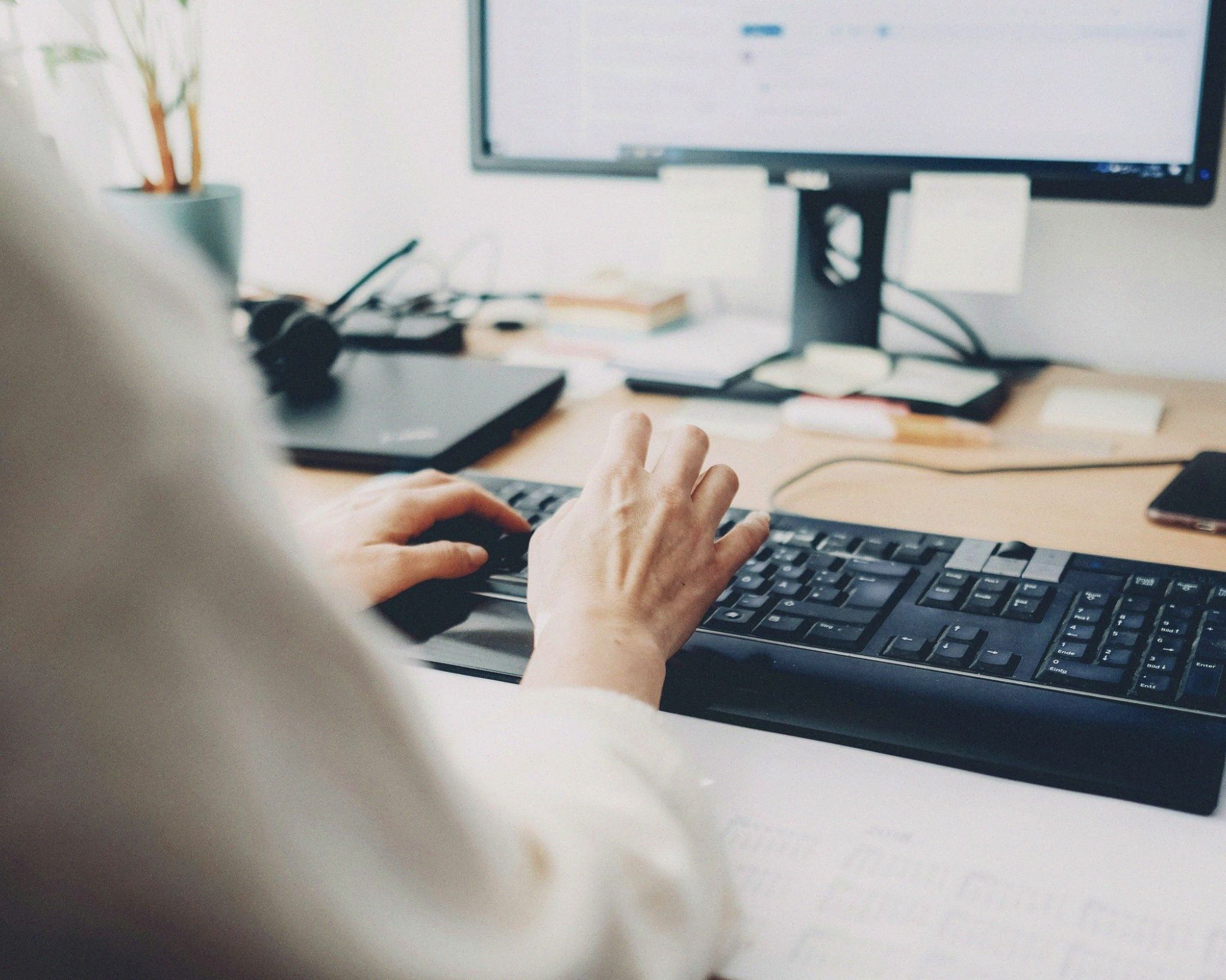 Person typing on a black keyboard at a desk with a computer monitor, laptop, and smartphone nearby.