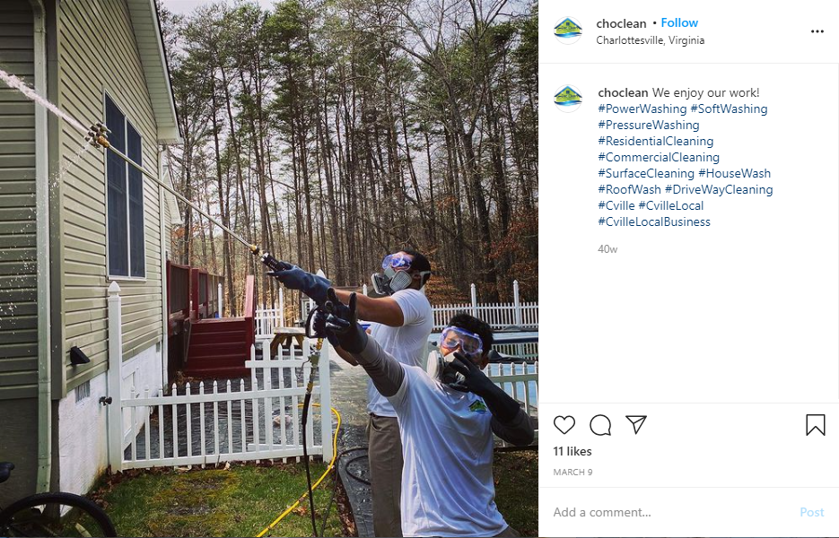 Workers in safety gear pressure washing house exterior near white fence