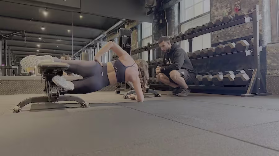 Female client performing glute-focused side plank with coach feedback at ONE|DZ Gym