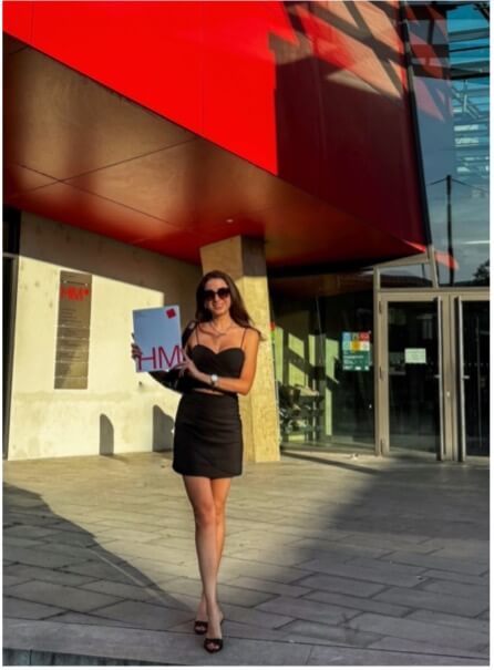 Жінка в чорній сукні тримає диплом перед сучасною будівлею з червоним фасадом, ймовірно, після церемонії вручення