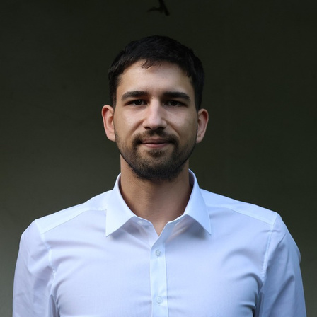 Portrait of a man with short dark hair and beard wearing a white collared shirt against a dark background.