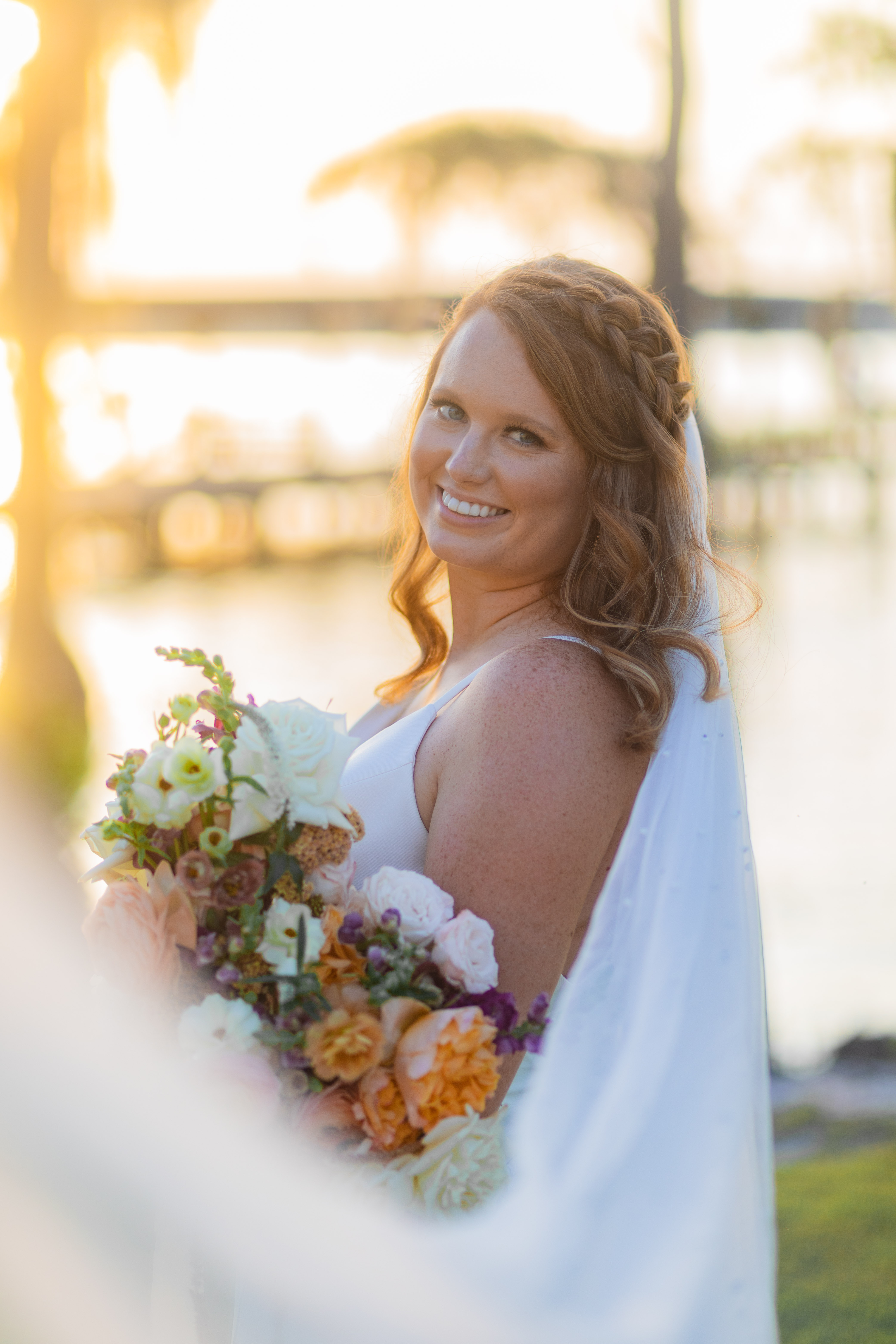 Bride in white dress smiling, holding colorful wedding bouquet at sunset