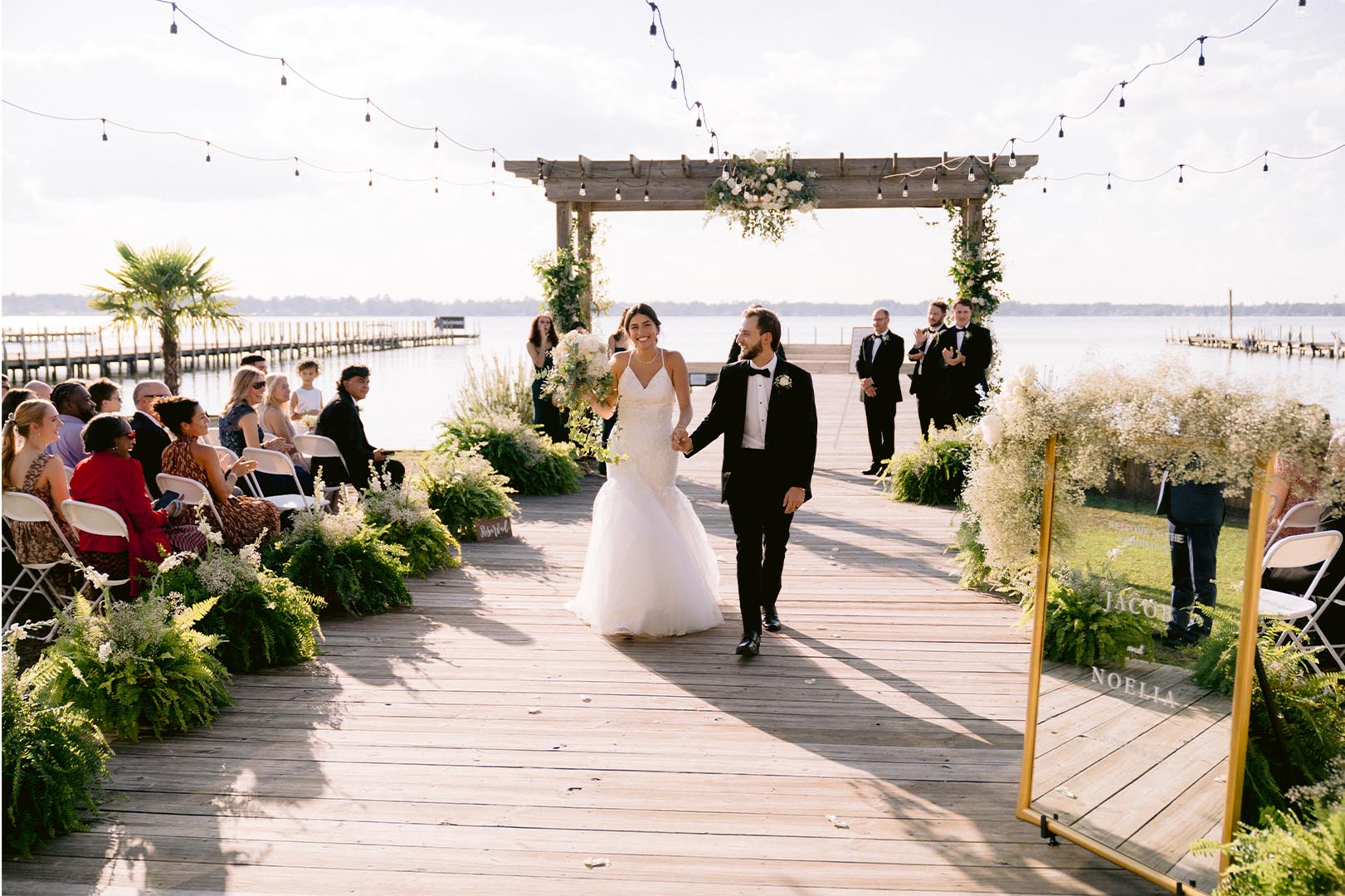 Waterfront wedding ceremony with wooden pier, string lights, and floral decorations