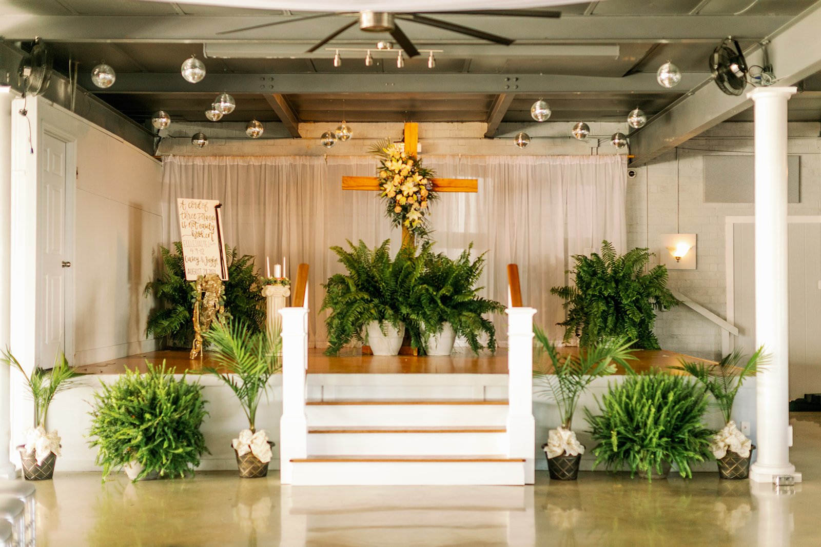 Wedding altar with yellow cross, white curtain, ferns, and hanging disco balls