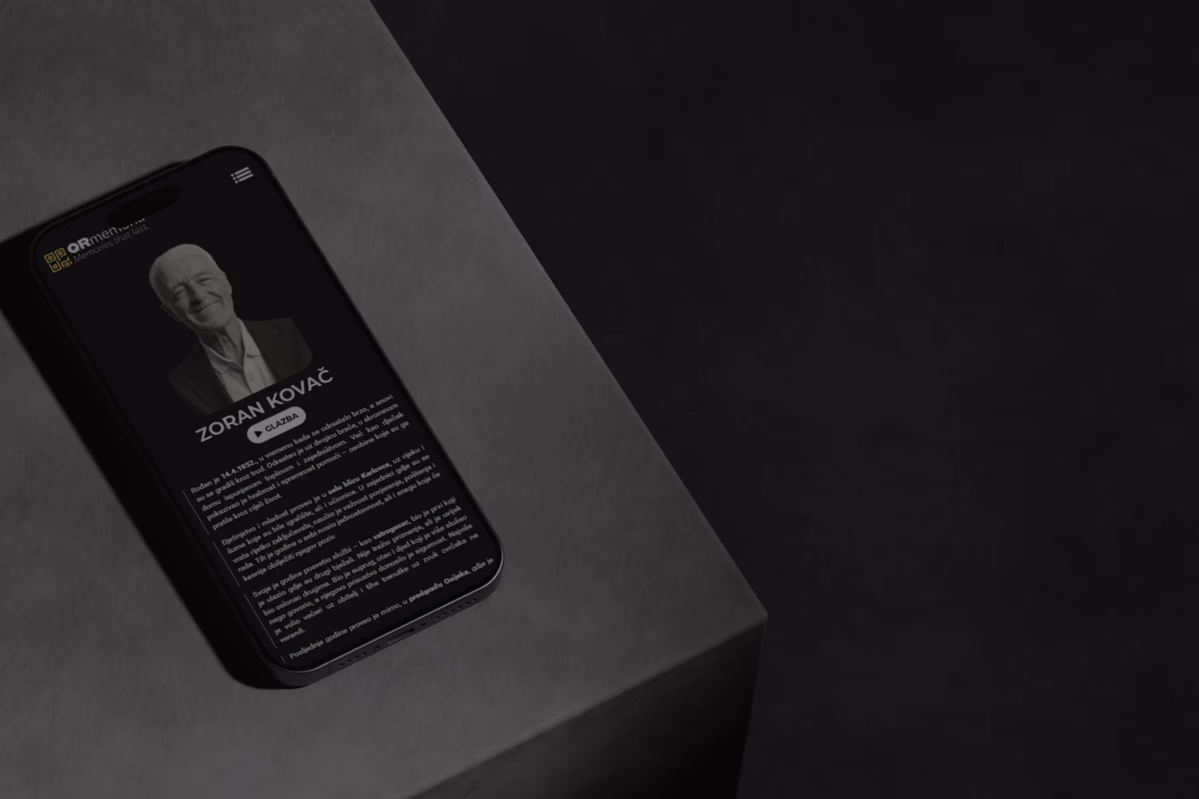 Pametni telefon na stolu prikazuje profil Zorana Kovača s crno-bijelom fotografijom i tekstom o njegovom životu.