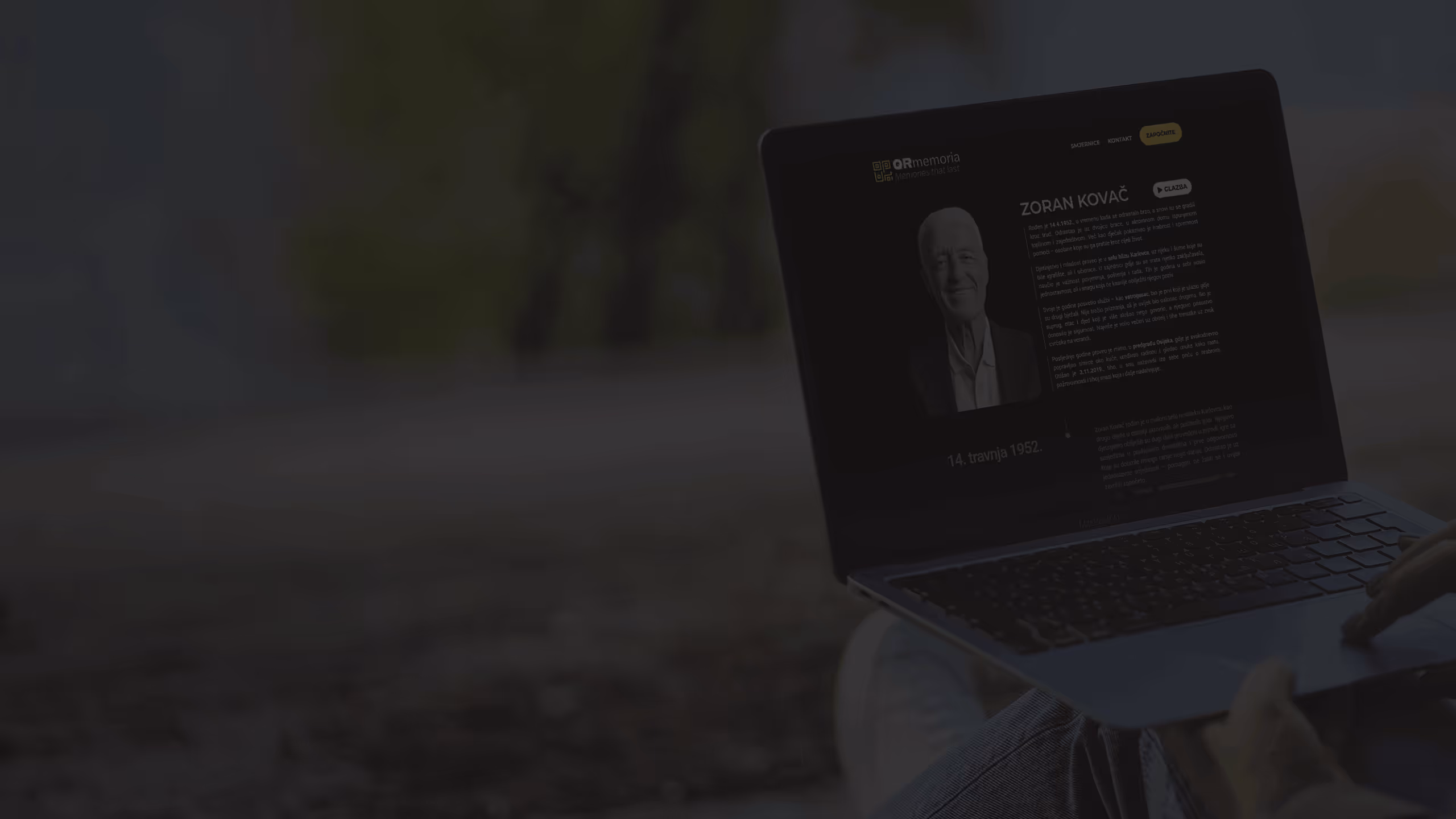 Osoba sjedi vani na tlu, tipka na prijenosnom računalu koje prikazuje stranicu s portretom i biografijom Zorana Kovača.