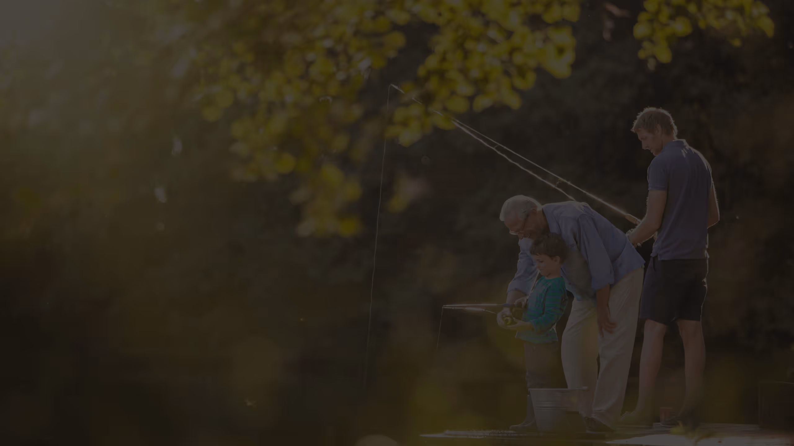 Tri generacije muškaraca stoje na pristaništu uz jezero, starac pomaže djetetu u ribolovu dok mlađi muškarac drži štap za ribolov.