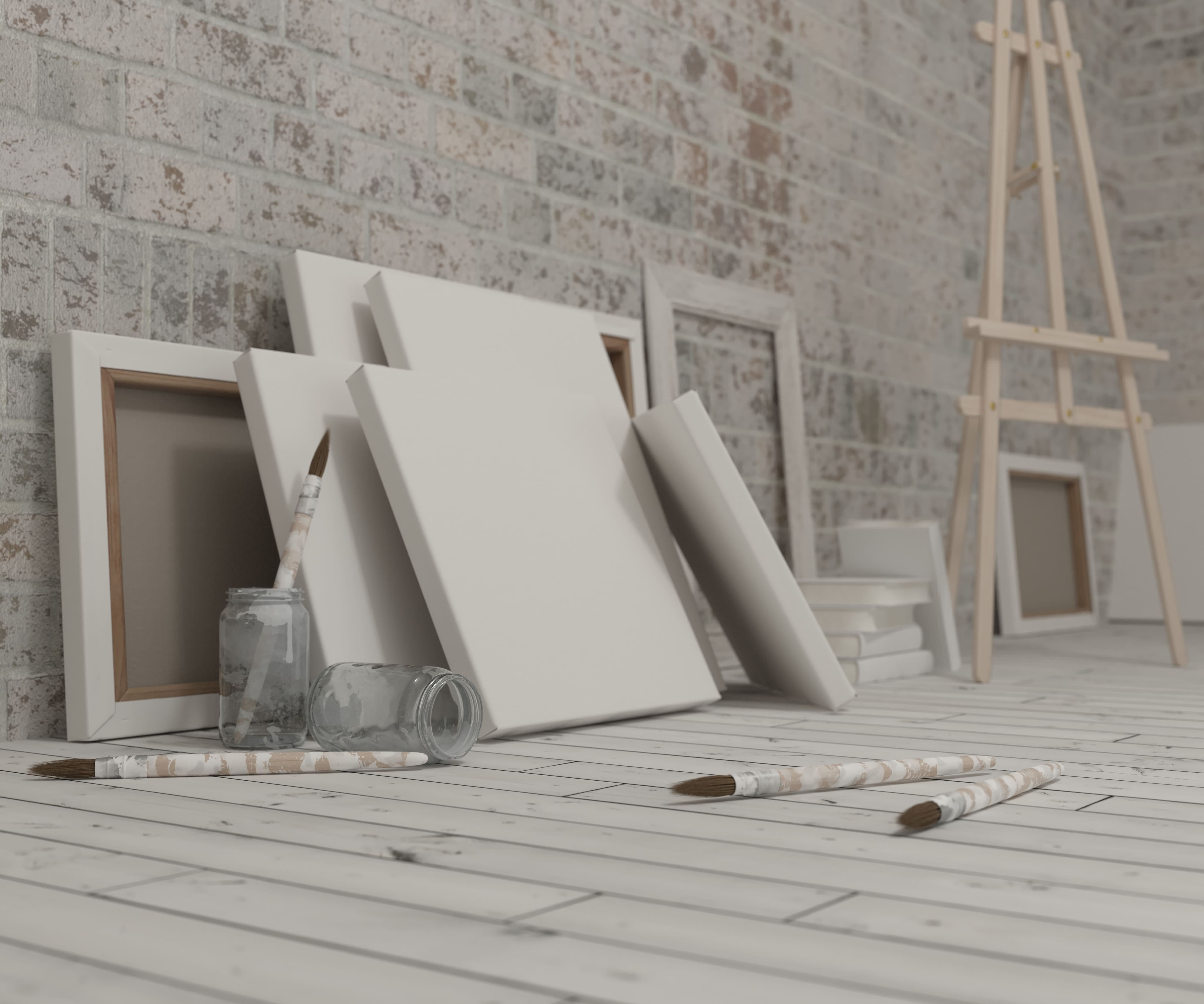 Multiple blank canvases leaning against a brick wall with paintbrushes and jars on a white wooden floor near a wooden easel.