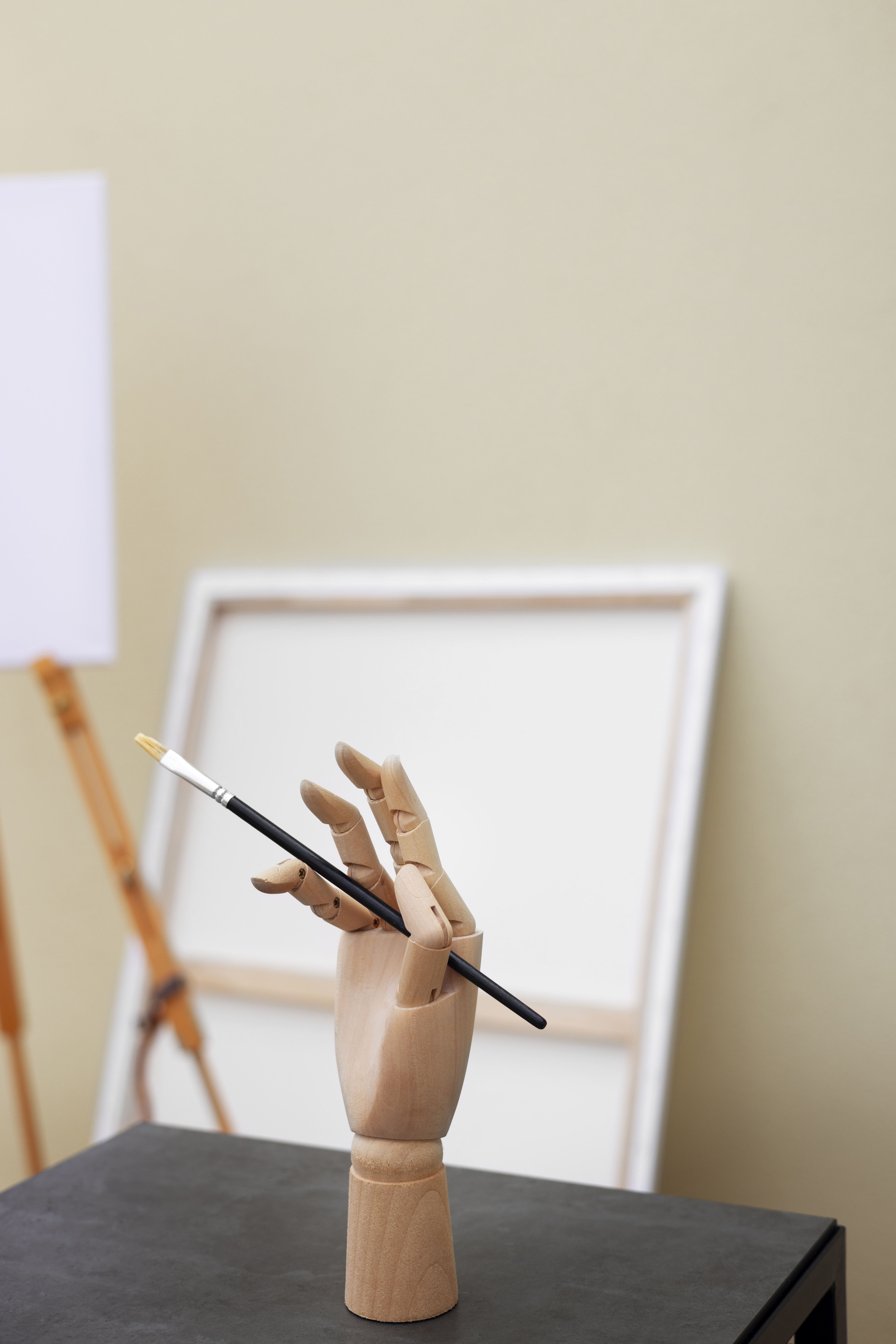 Wooden artist's mannequin hand holding a paintbrush upright, with a blurred blank canvas and easel in the background.