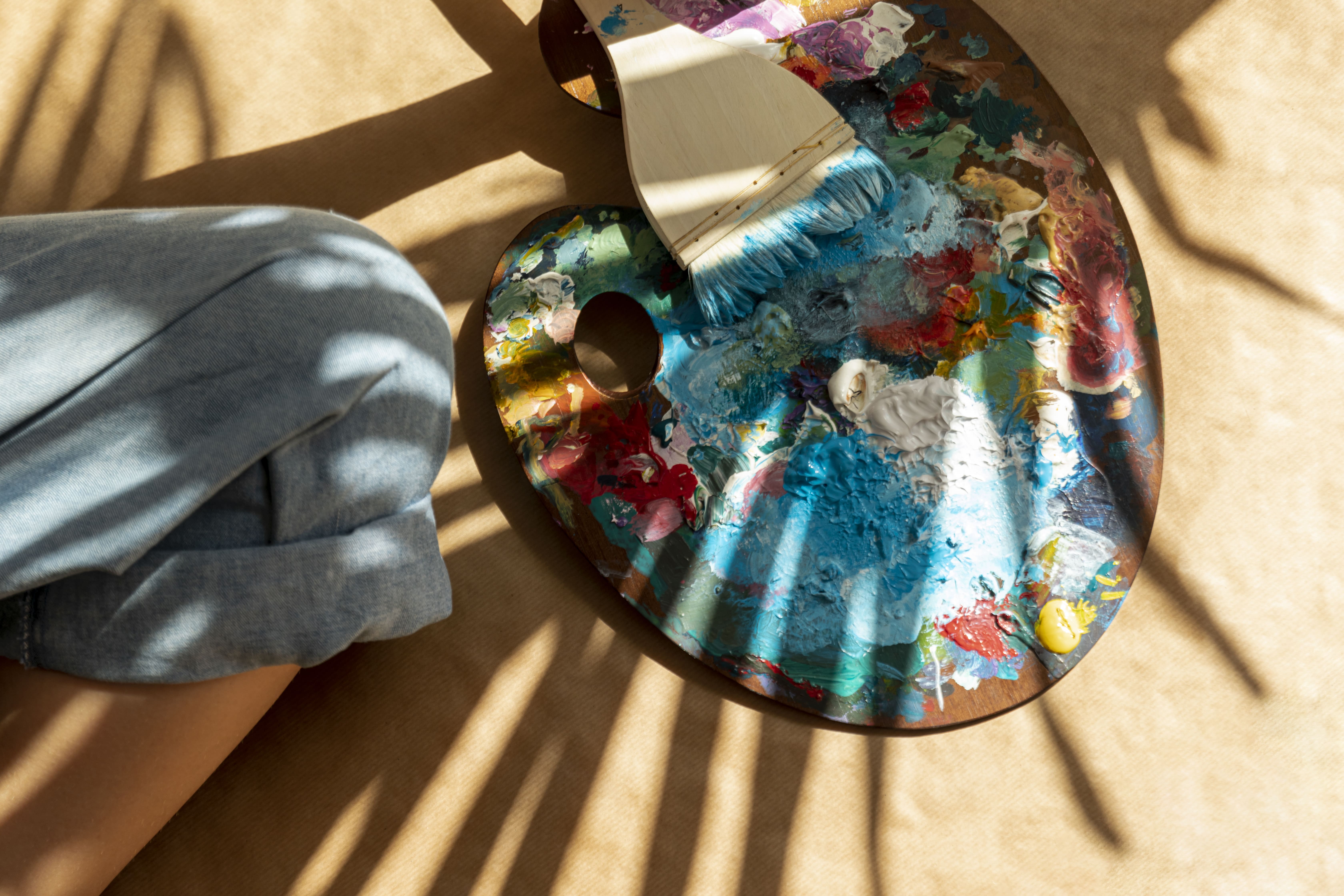 Artist's paint palette with various mixed colors and a paintbrush with blue paint resting on a beige surface, next to a person wearing blue jeans.