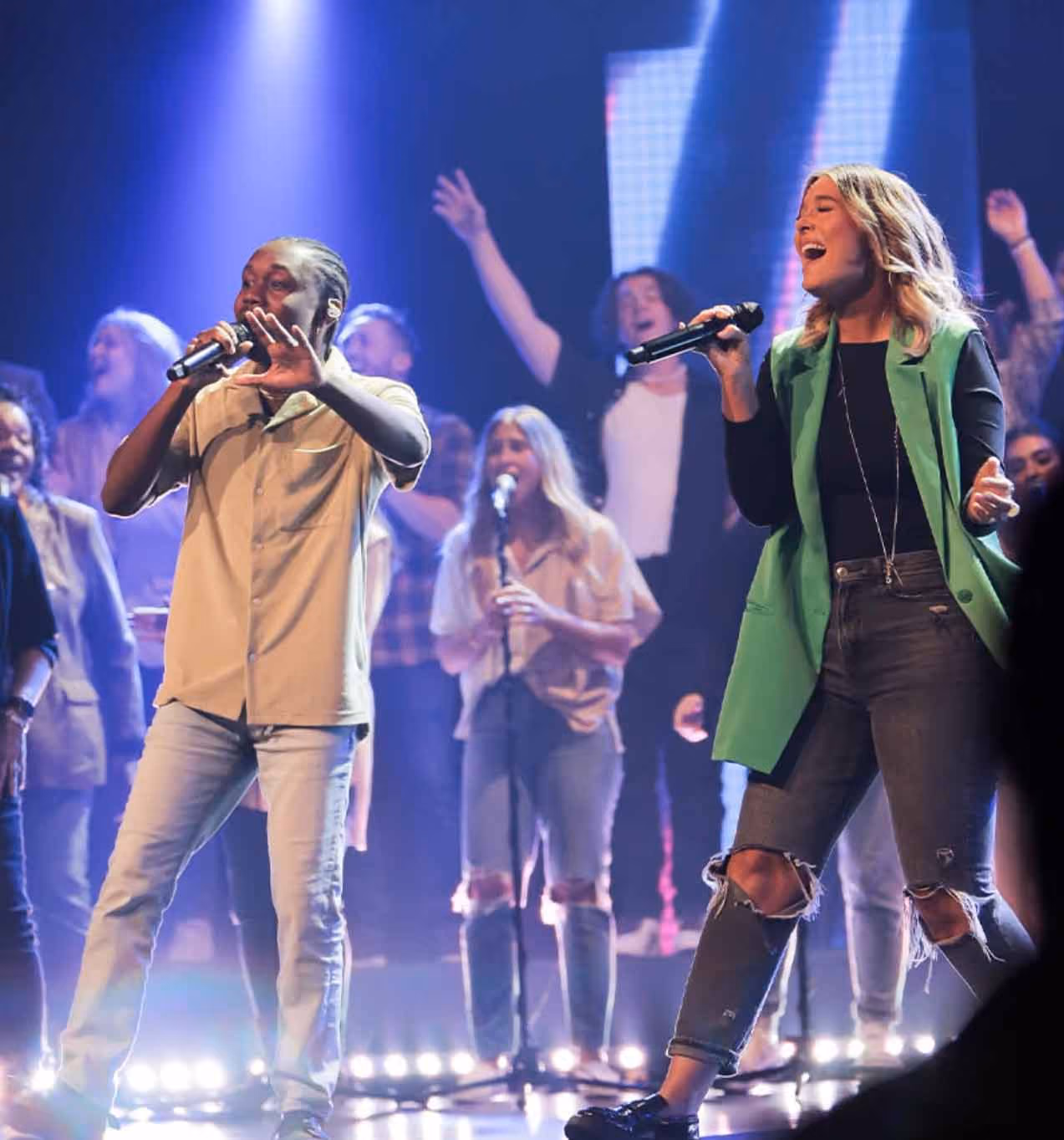 A diverse group of singers performing on stage with two lead vocalists holding microphones, one wearing a green vest and the other a beige shirt, illuminated by stage lights and accompanied by background singers.