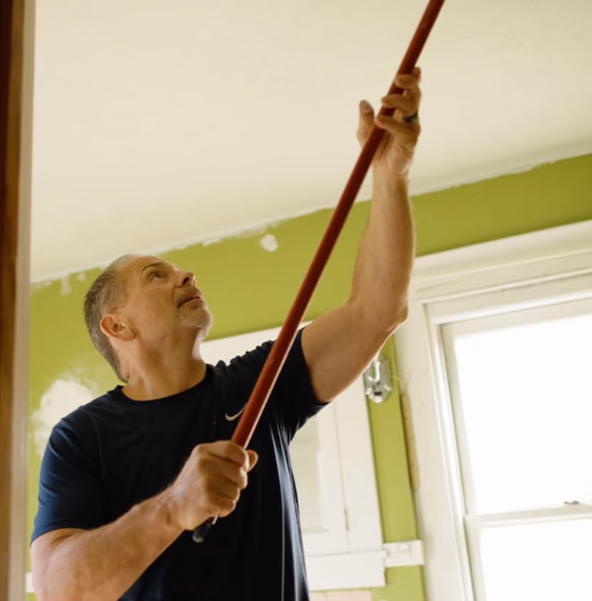Man in a dark shirt painting or rolling a ceiling white using a long-handled paint roller in a room with green walls and a window.