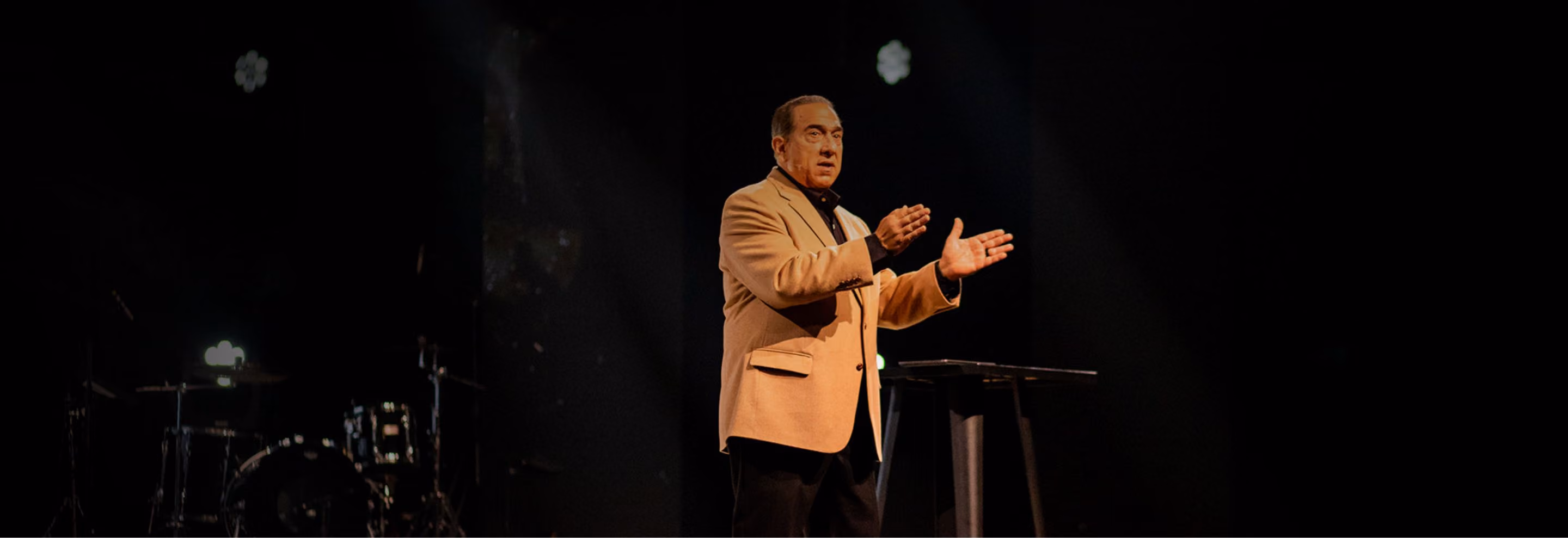 Man in a brown blazer speaking on a dark stage with a spotlight, standing near a podium with drums visible in the background.