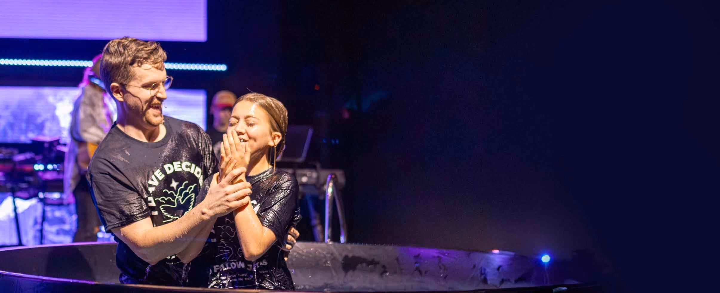 Man baptizing woman in a pool during a religious ceremony, both smiling and wearing dark shirts with a logo, with stage and musical equipment in the background.