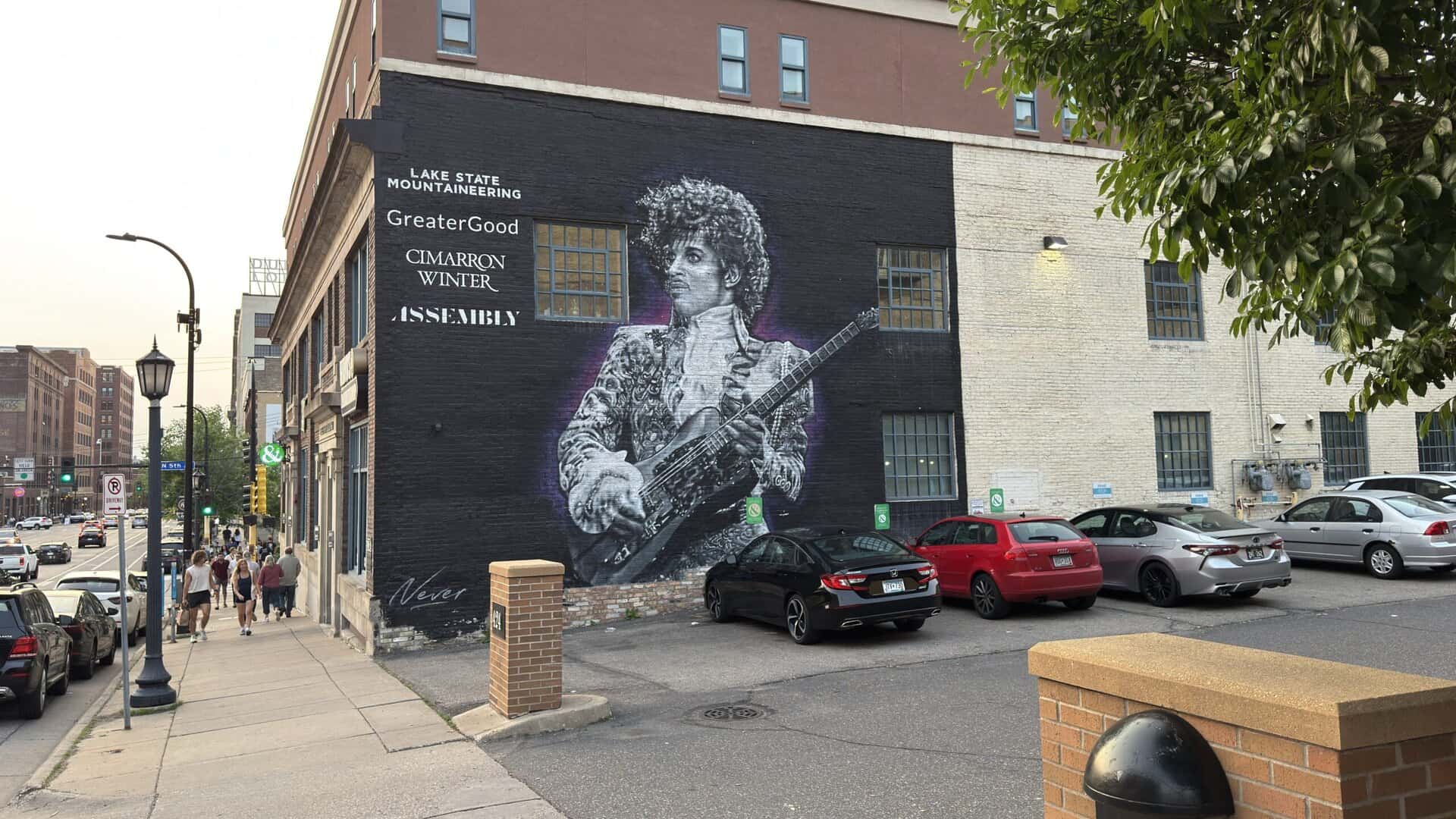 Street view of Prince Mural in North Loop Minneapolis painted on a building wall playing guitar, surrounded by sponsor names, with parked cars and pedestrians nearby.