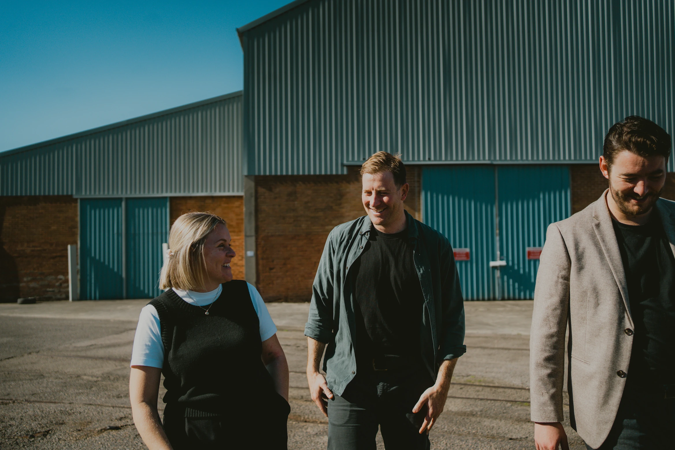Three people walking together with tin industrial shed behind them