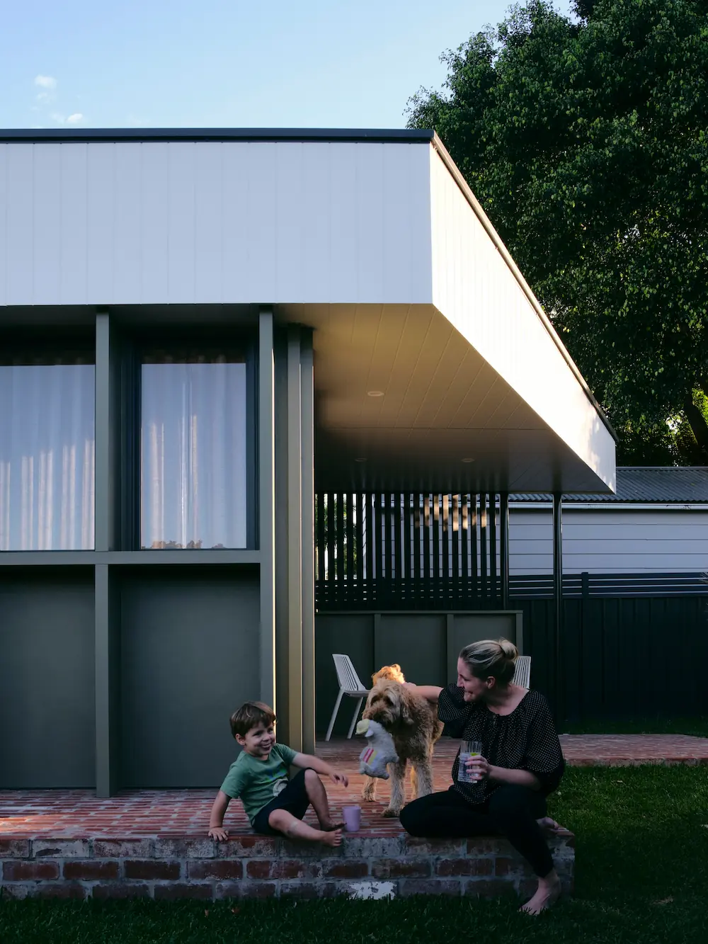 Mother and son sit on porch with their dog with modern house in background