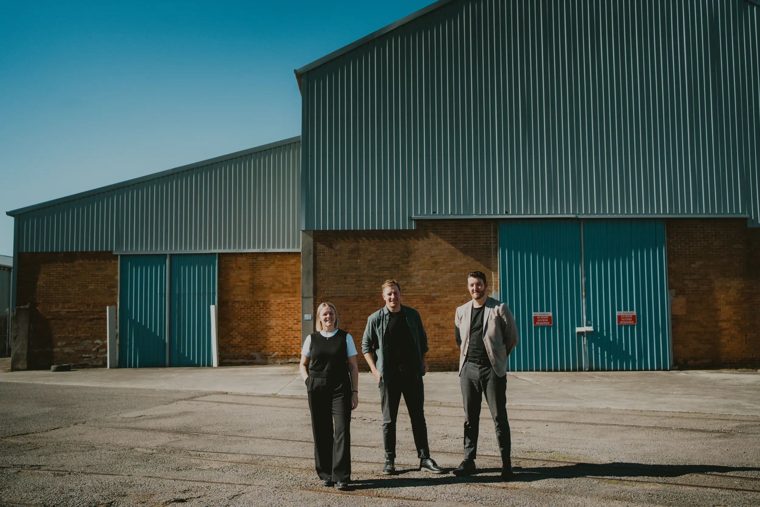 Three people standing in front of brick and tin warehouse looking at camera