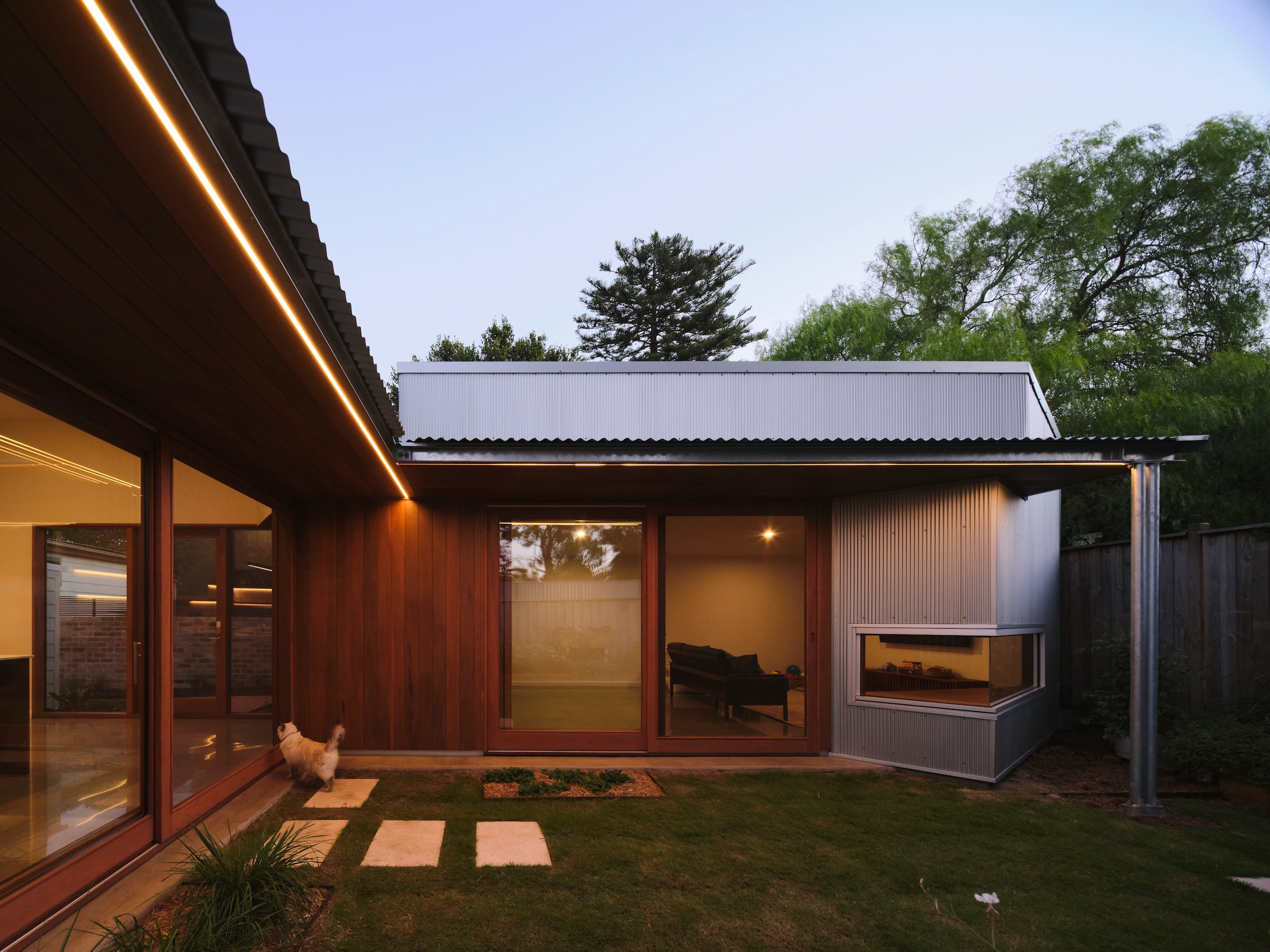 Modern single-story house exterior at dusk with wood and metal siding, large windows, and a cat near the glass door.