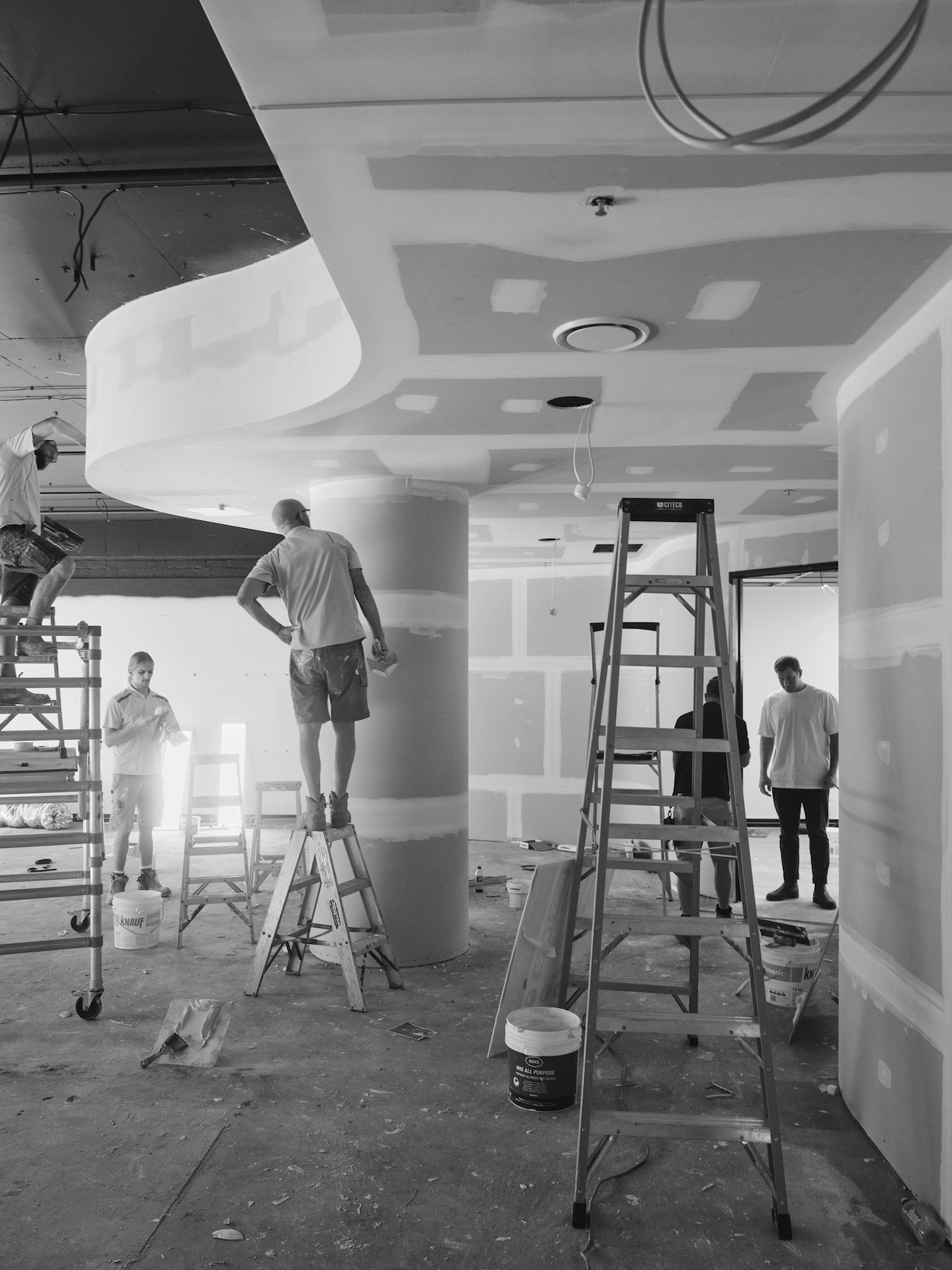 Construction workers performing drywall and plasterwork inside a building under renovation with ladders and tools scattered on the floor.