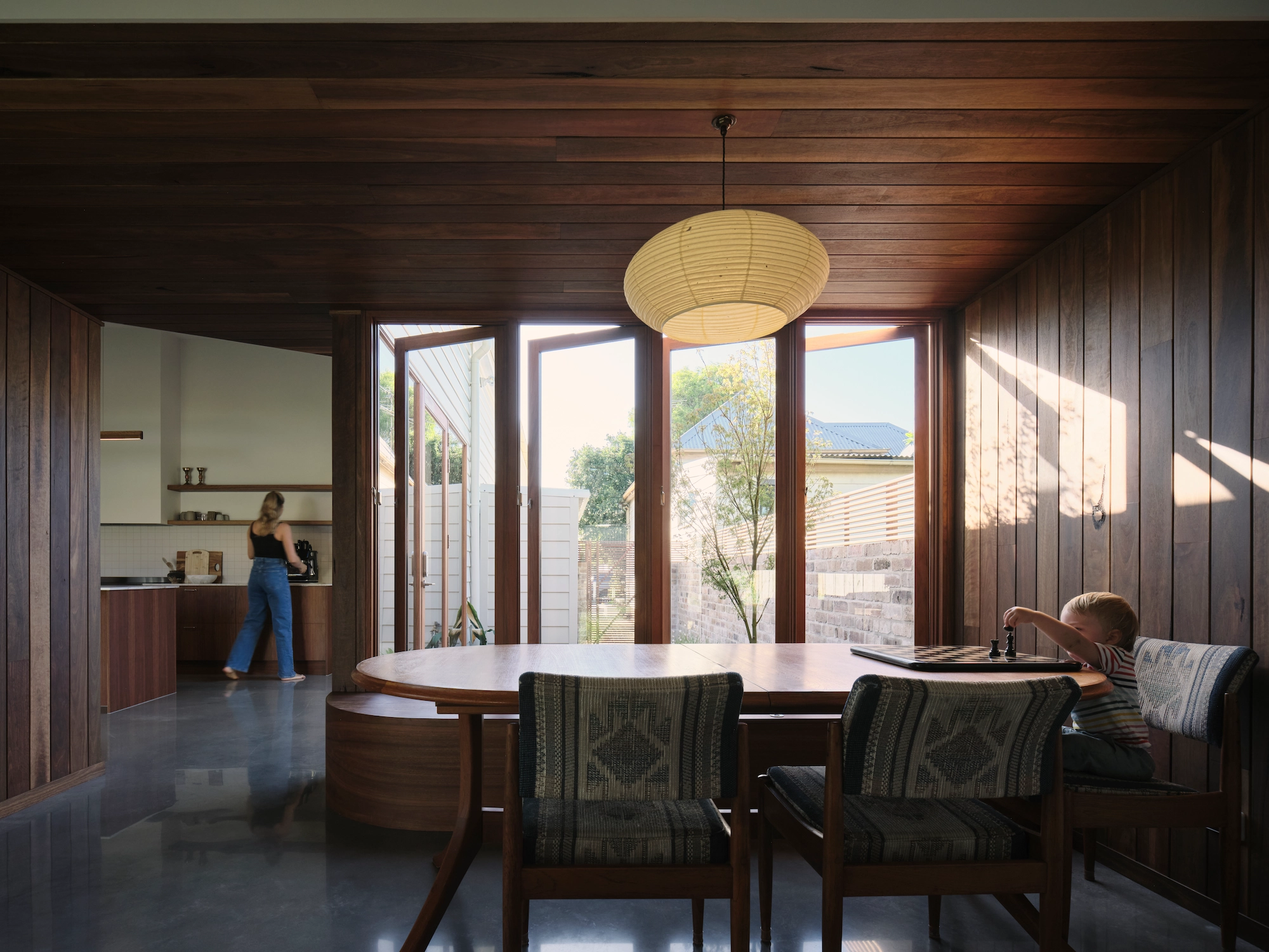 Modern dining room with wooden walls and ceiling, large glass doors leading outside, a round wooden table with chairs, and a child playing chess on the right side.