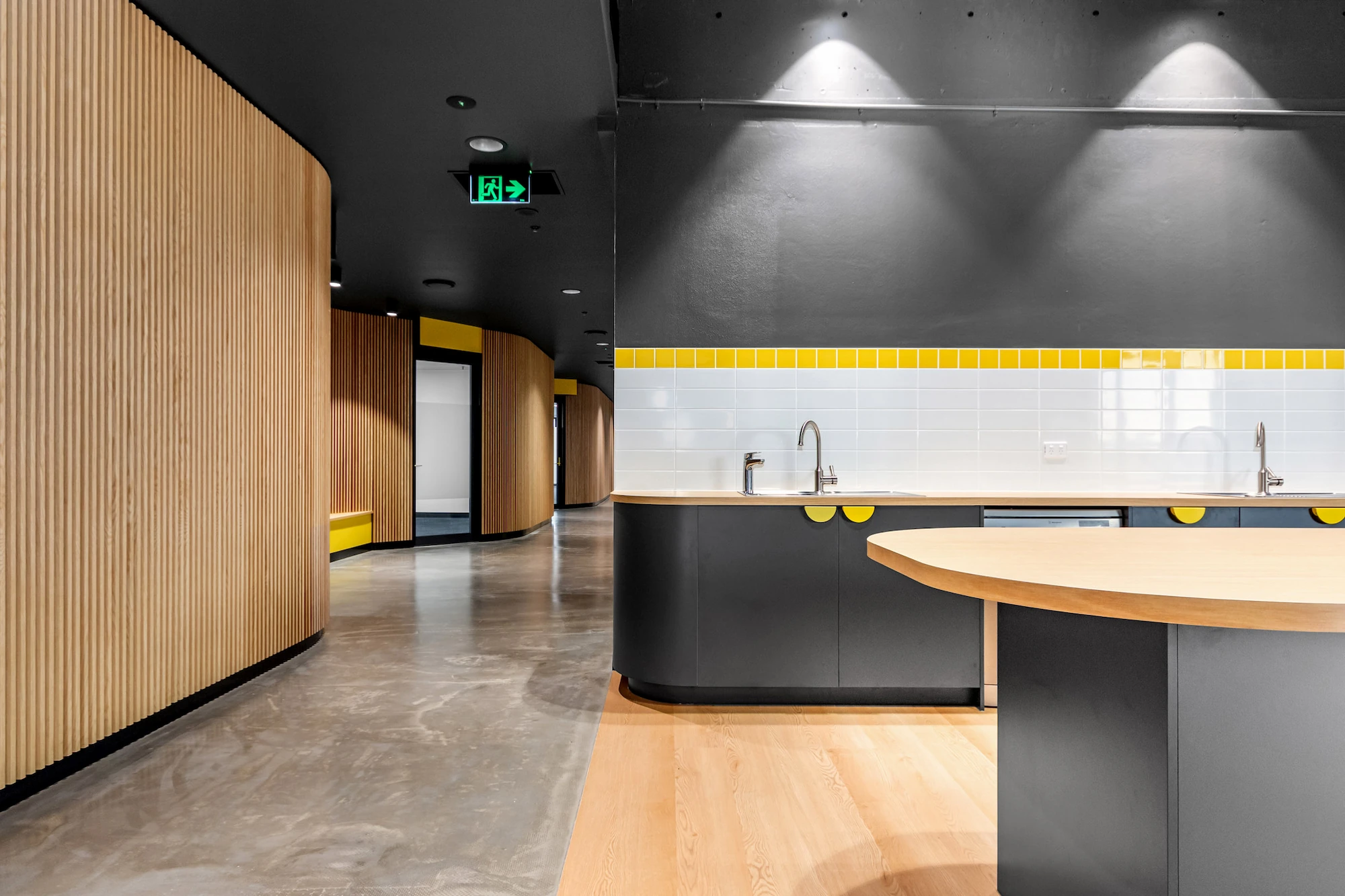 Modern kitchen area with black cabinets, wooden countertops, white tiled backsplash with yellow trim, and curved wooden wall panels beside a polished concrete floor.