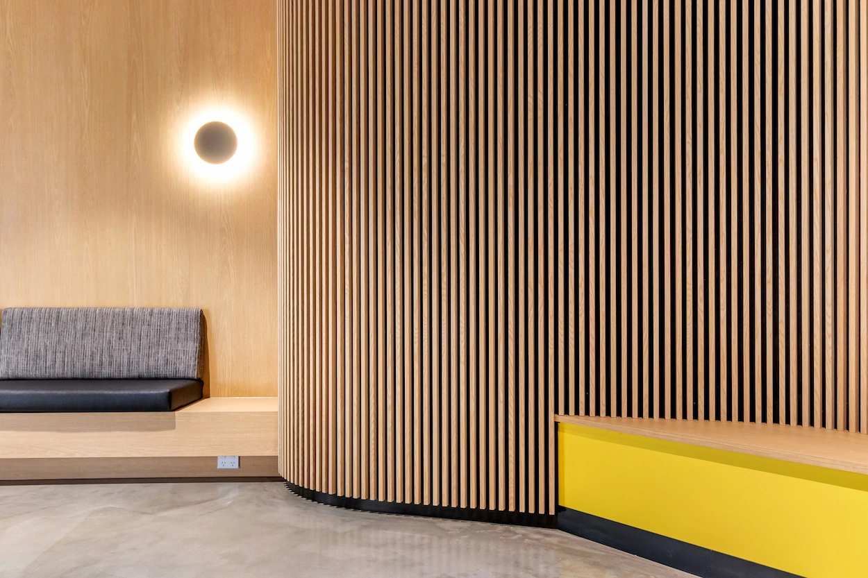 Modern interior with curved vertical wooden slats, a gray cushioned bench, yellow accent seating, and a circular wall light.