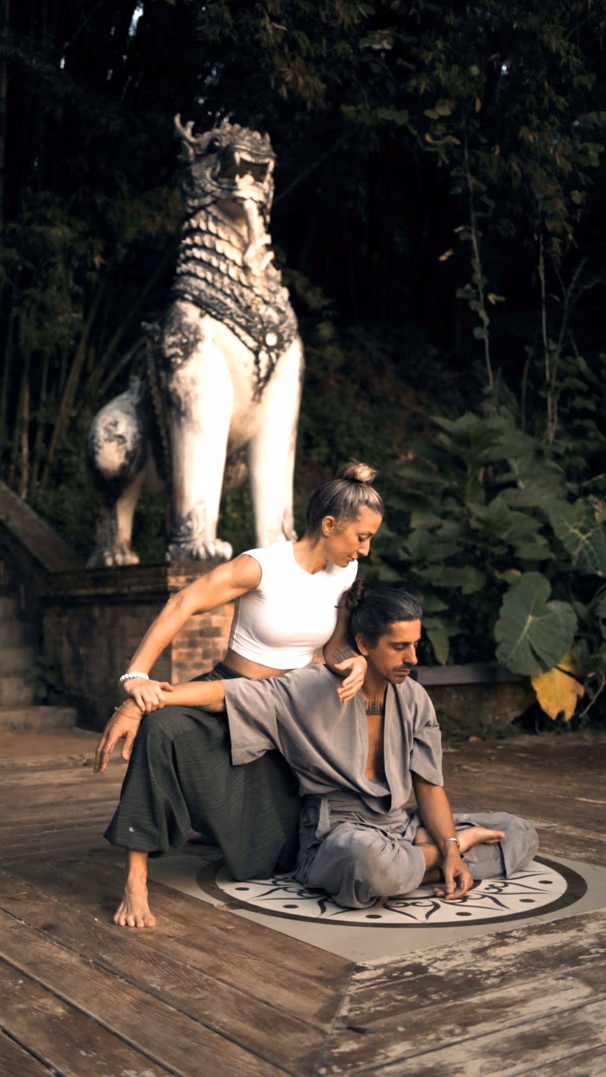 Two people practicing yoga near stone lion statue in lush garden