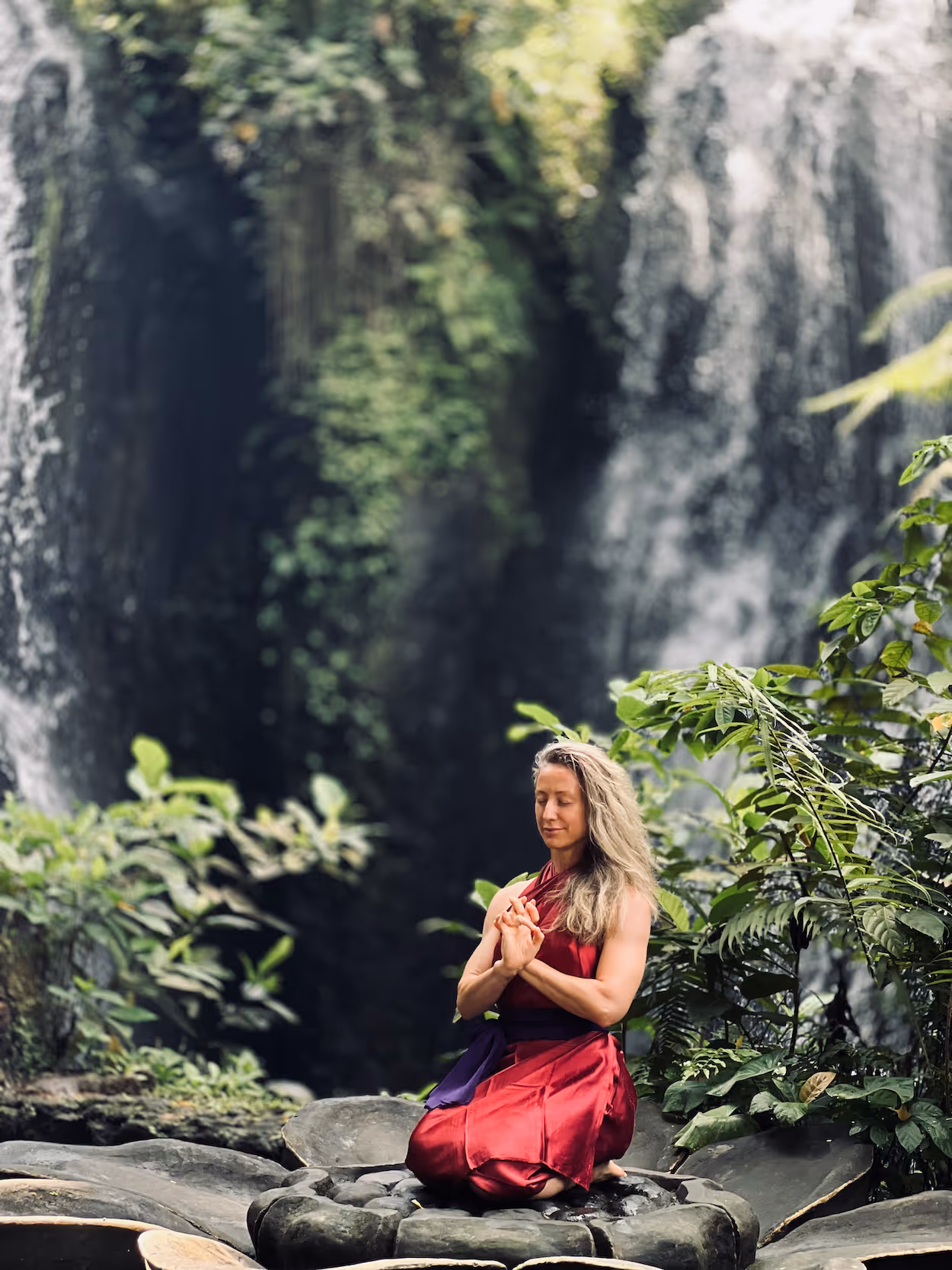 Woman meditating in lotus position near waterfall surrounded by lush greenery