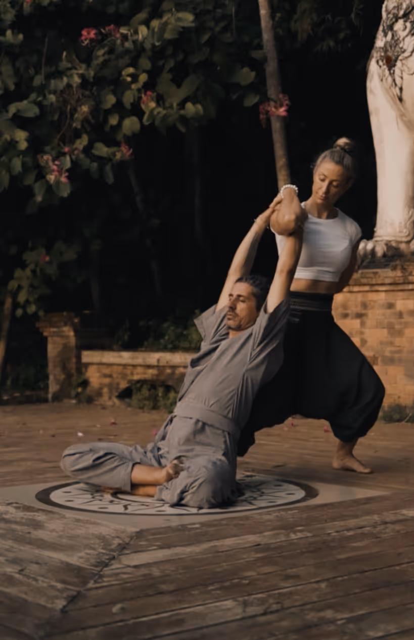 Two people practicing yoga on wooden platform with flowering plants background