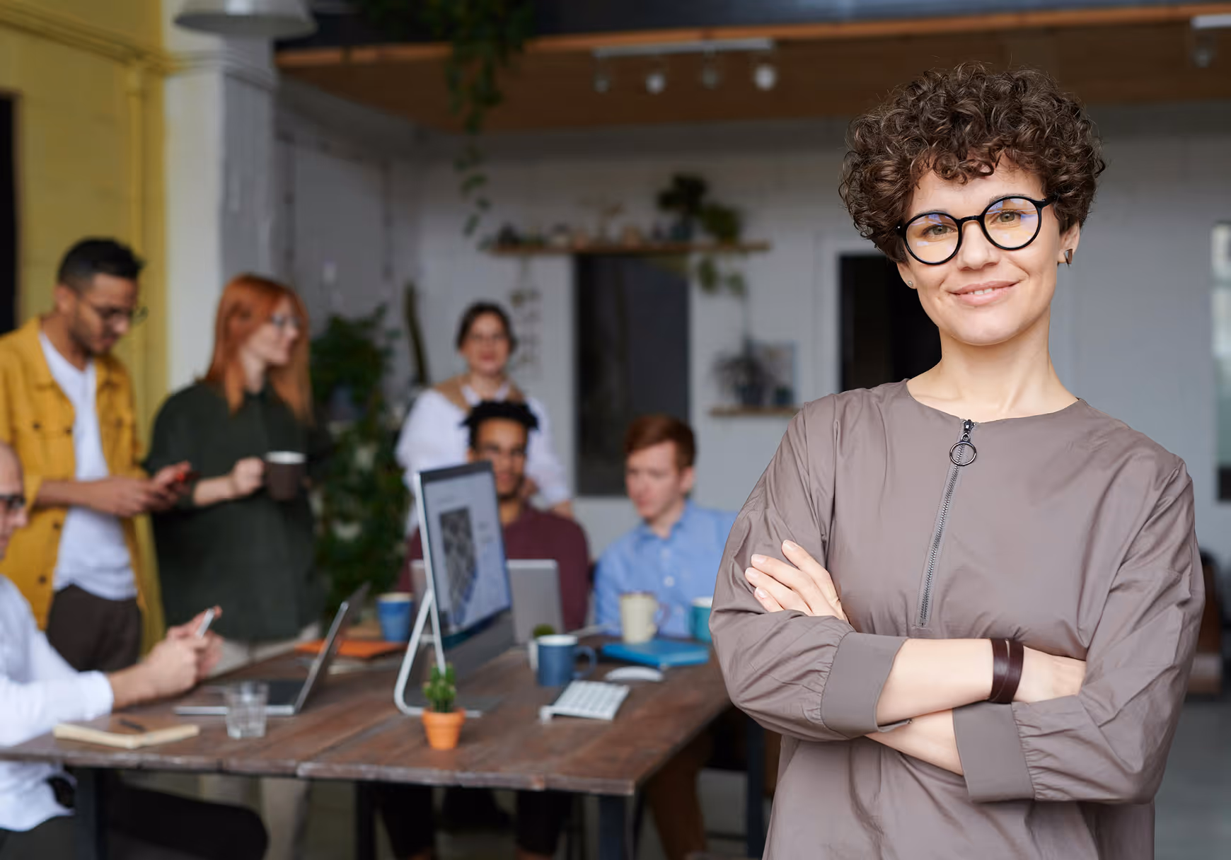 Confident woman with curly hair and glasses standing with crossed arms in a modern office with a group of colleagues working in the background.