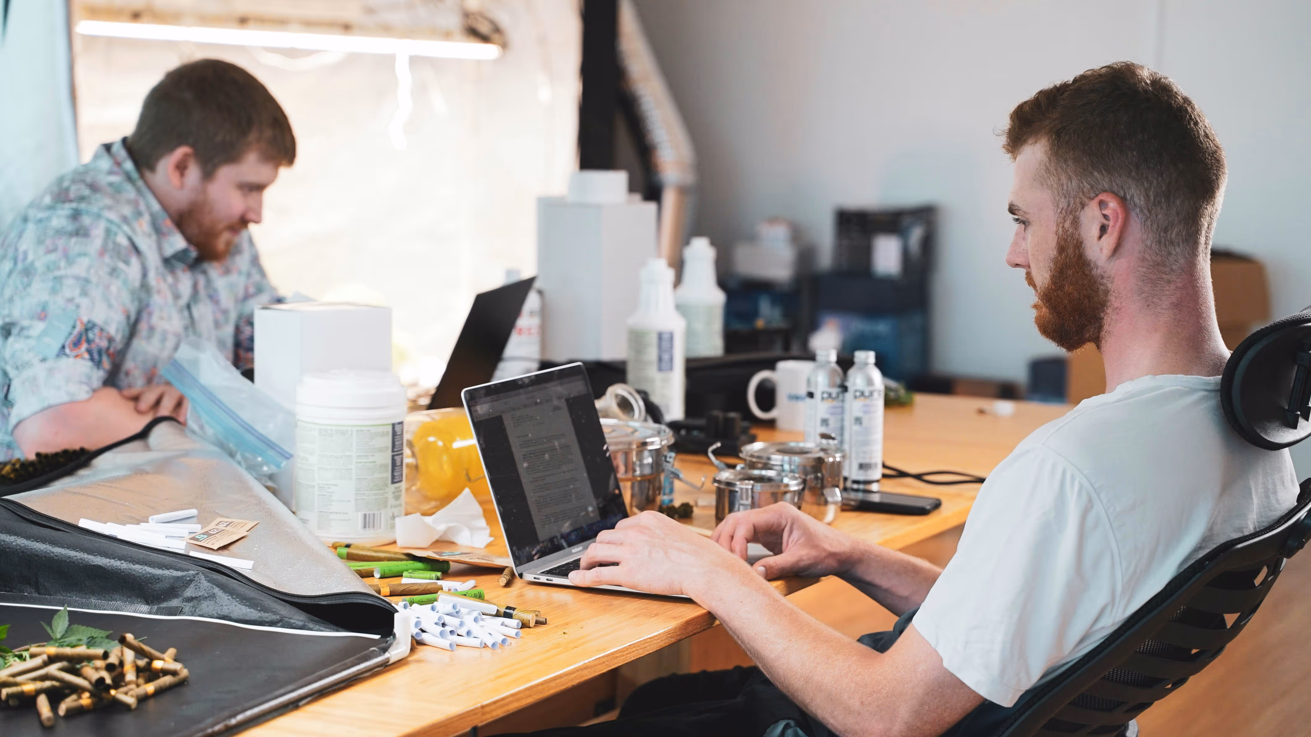 Two men working on laptops at a desk cluttered with various items including containers, bottles, and small plant parts.