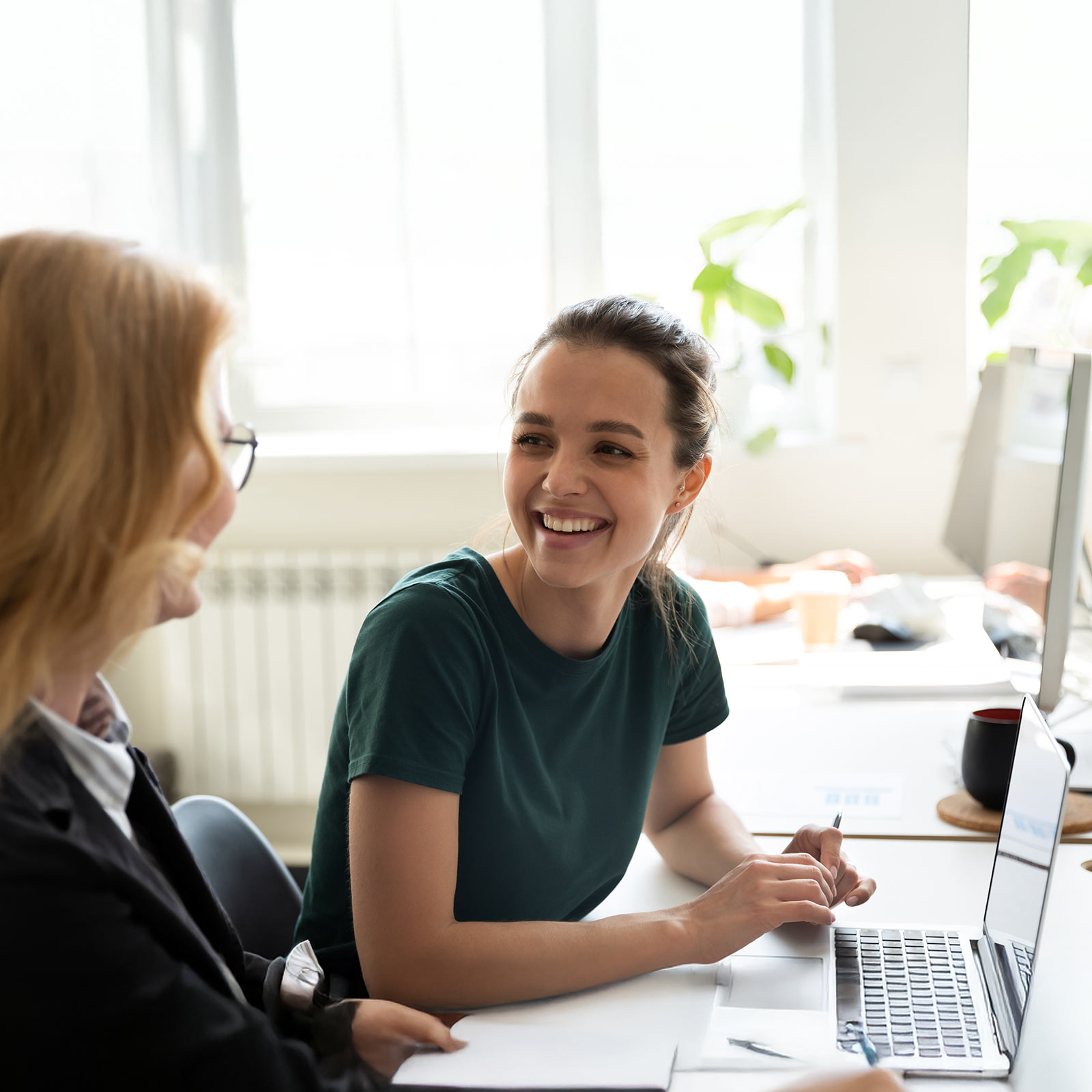Two woman sitting at a desk in front of a laptop smiling and talking
