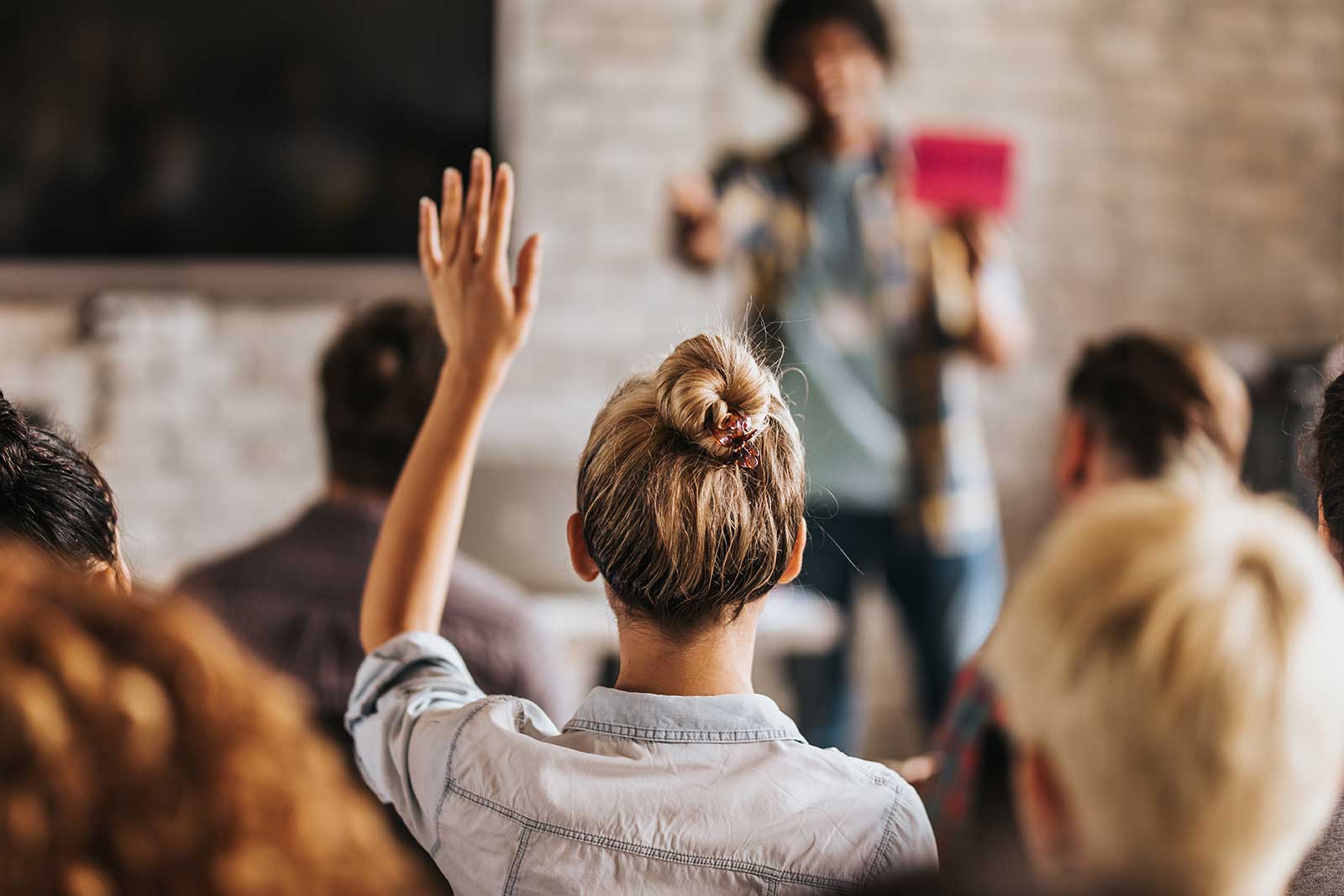 A woman seated in room of people, with her hand up to ask a question