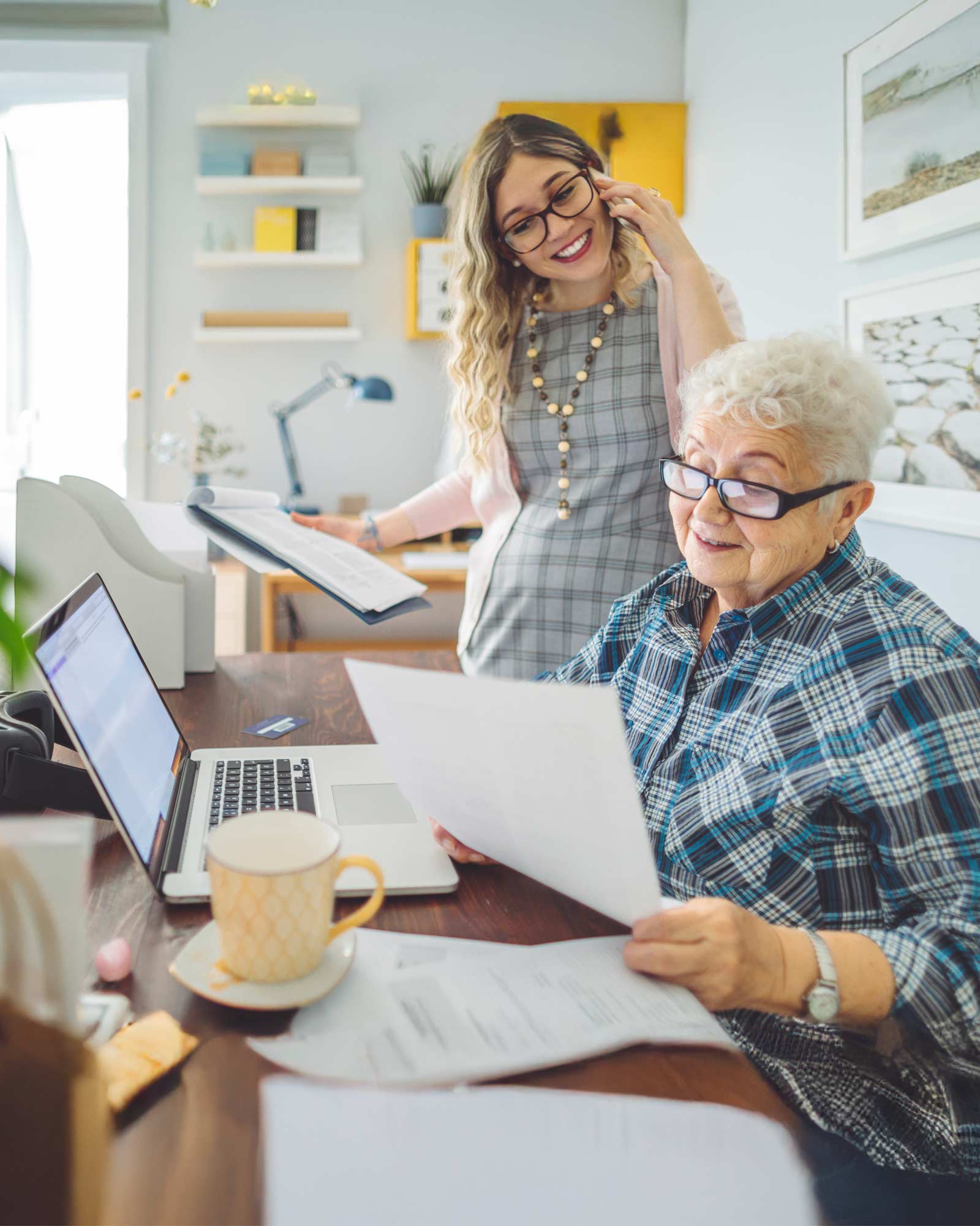 An older woman seated at a desk looking at some paperwork in front of her laptop, with another woman on the phone, smiling, looking on.